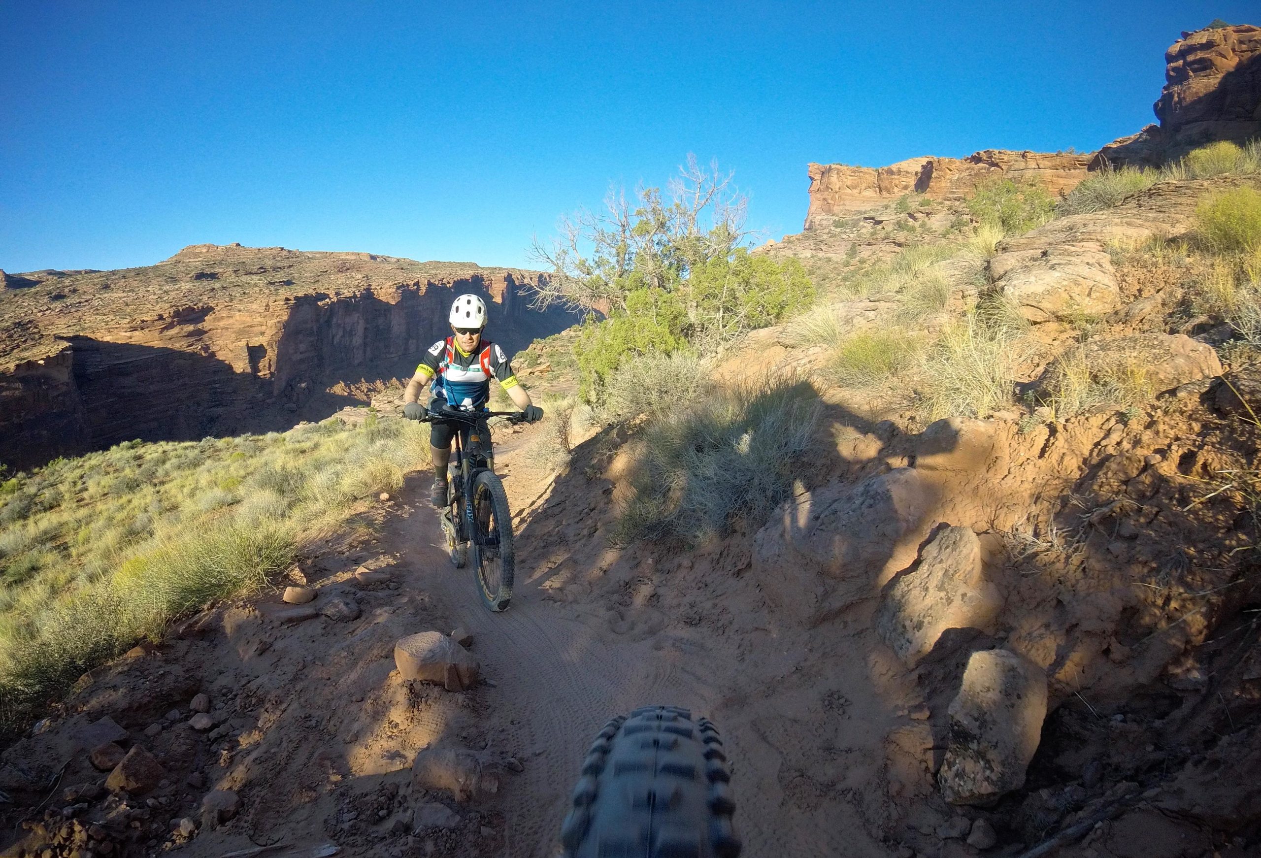 A mountain biker riding along a dusty trail with rocky terrain and desert vegetation, against the backdrop of cliffs and a clear blue sky. The angle suggests a view from another rider’s perspective, showcasing the rider’s helmet and bike. Porcupine Rim mountain bike trail.