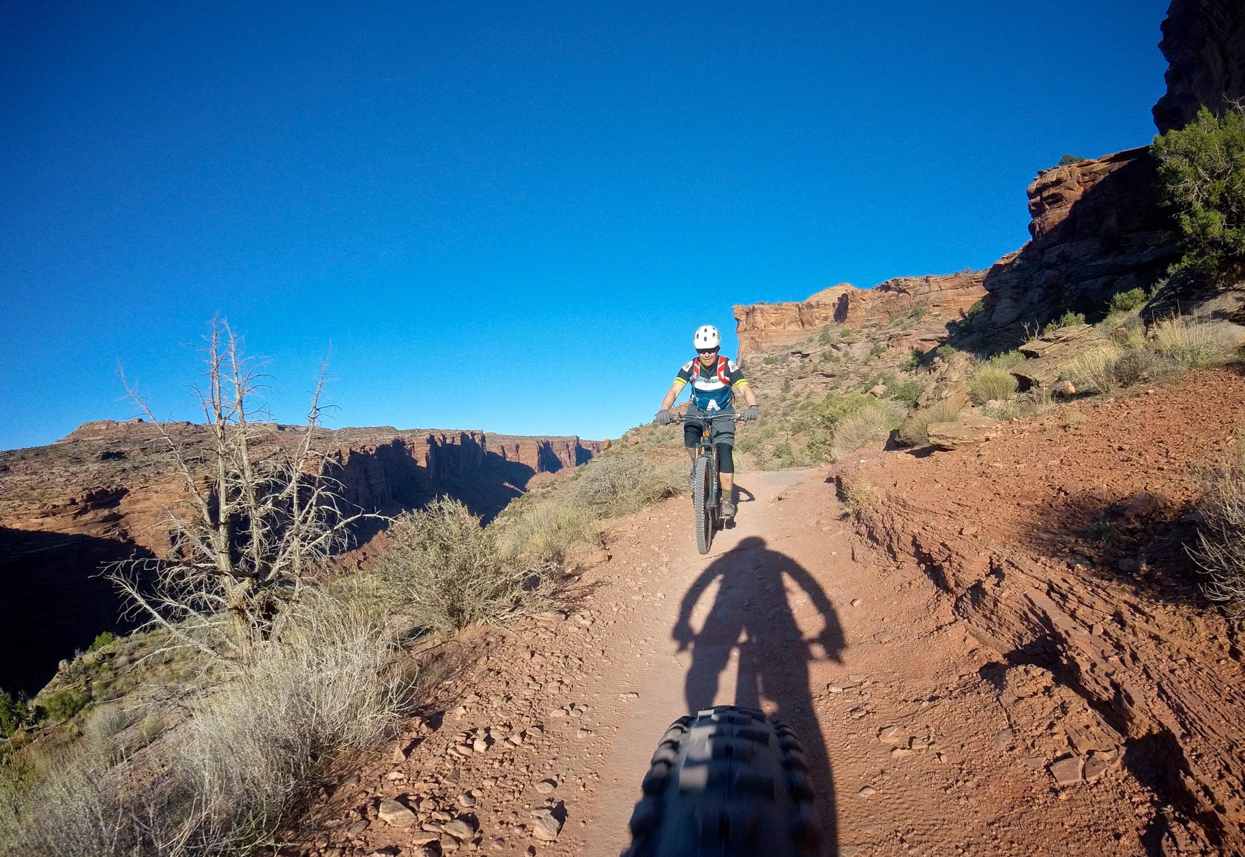 A mountain biker riding along a dirt trail with a clear blue sky and rocky terrain in the background. The biker is wearing a white helmet and colorful cycling gear, and their shadow is visible on the ground. Surrounding vegetation includes sparse shrubs and a dry, leafless tree. Porcupine Rim mountain bike trail.
