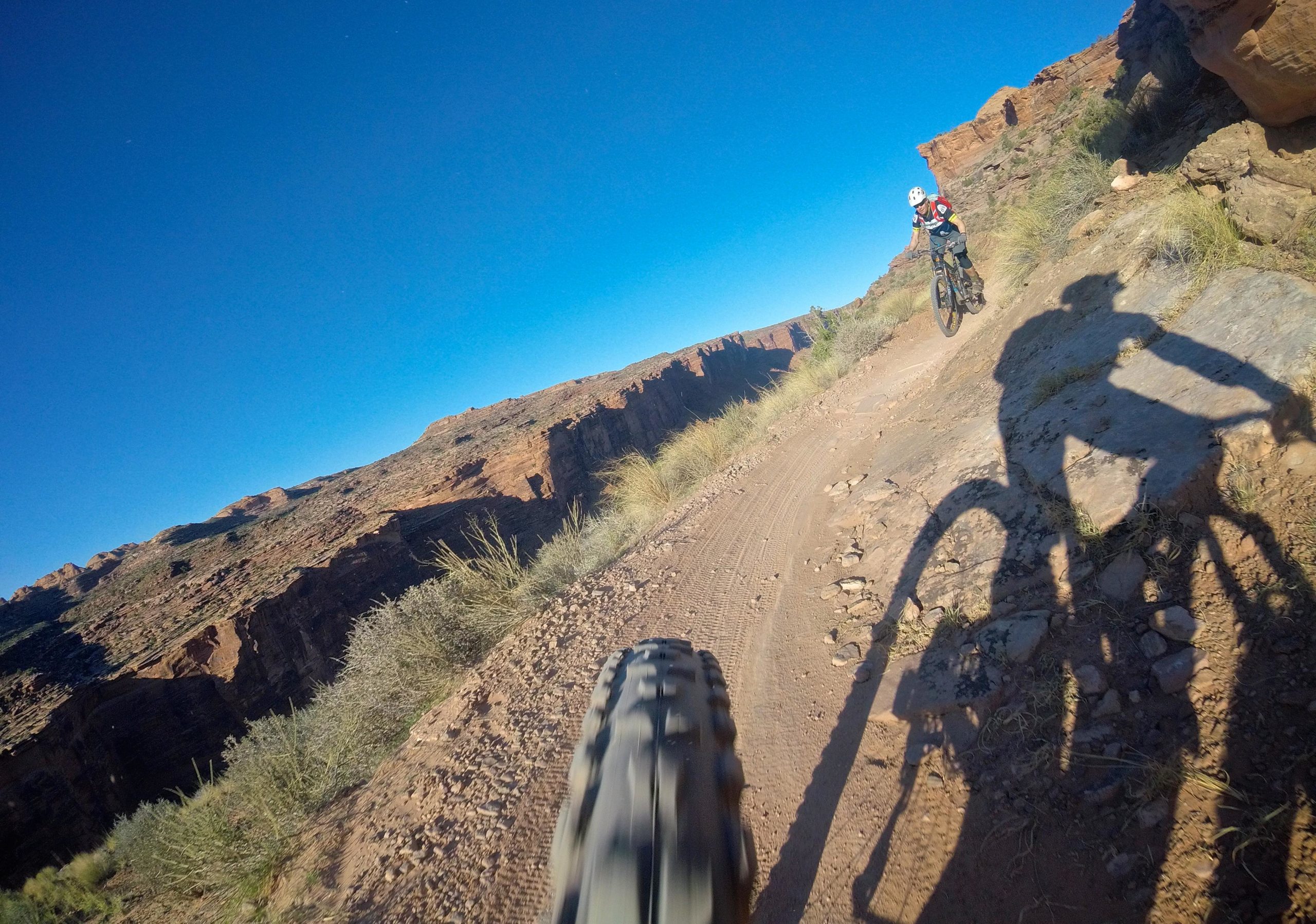 A mountain biker rides along a narrow dirt trail with rocky terrain, surrounded by steep canyon walls under a clear blue sky. The photo captures the biker's shadow and the bike's tire in the foreground, emphasizing the adventurous nature of the ride. Porcupine Rim mountain bike trail.