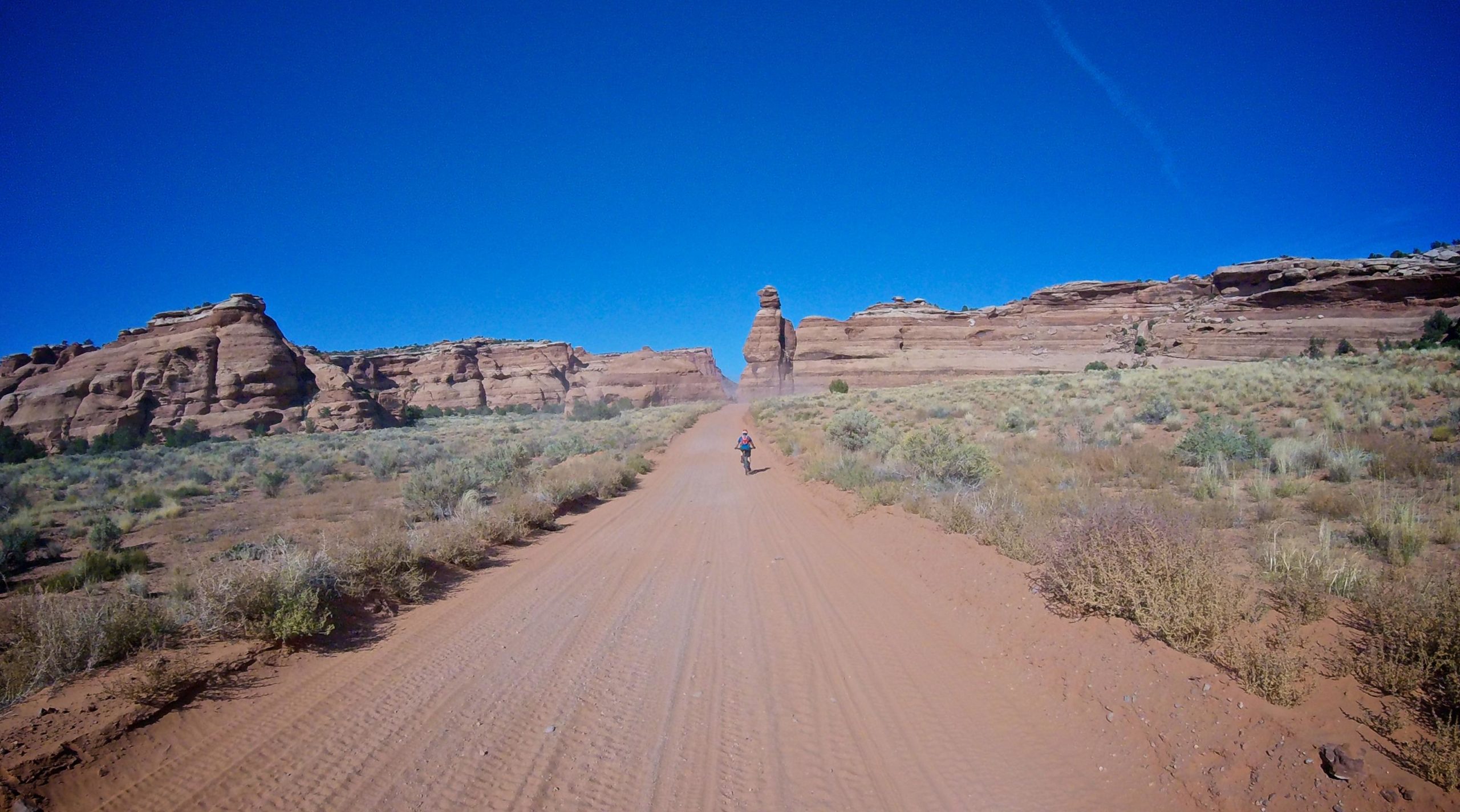 A person riding a bicycle along a dirt path in a desert landscape, surrounded by rock formations under a clear blue sky. The path leads toward a prominent rock spire in the distance, with sparse vegetation and the texture of the sandy ground visible. Gemini Bridges mountain bike trail.