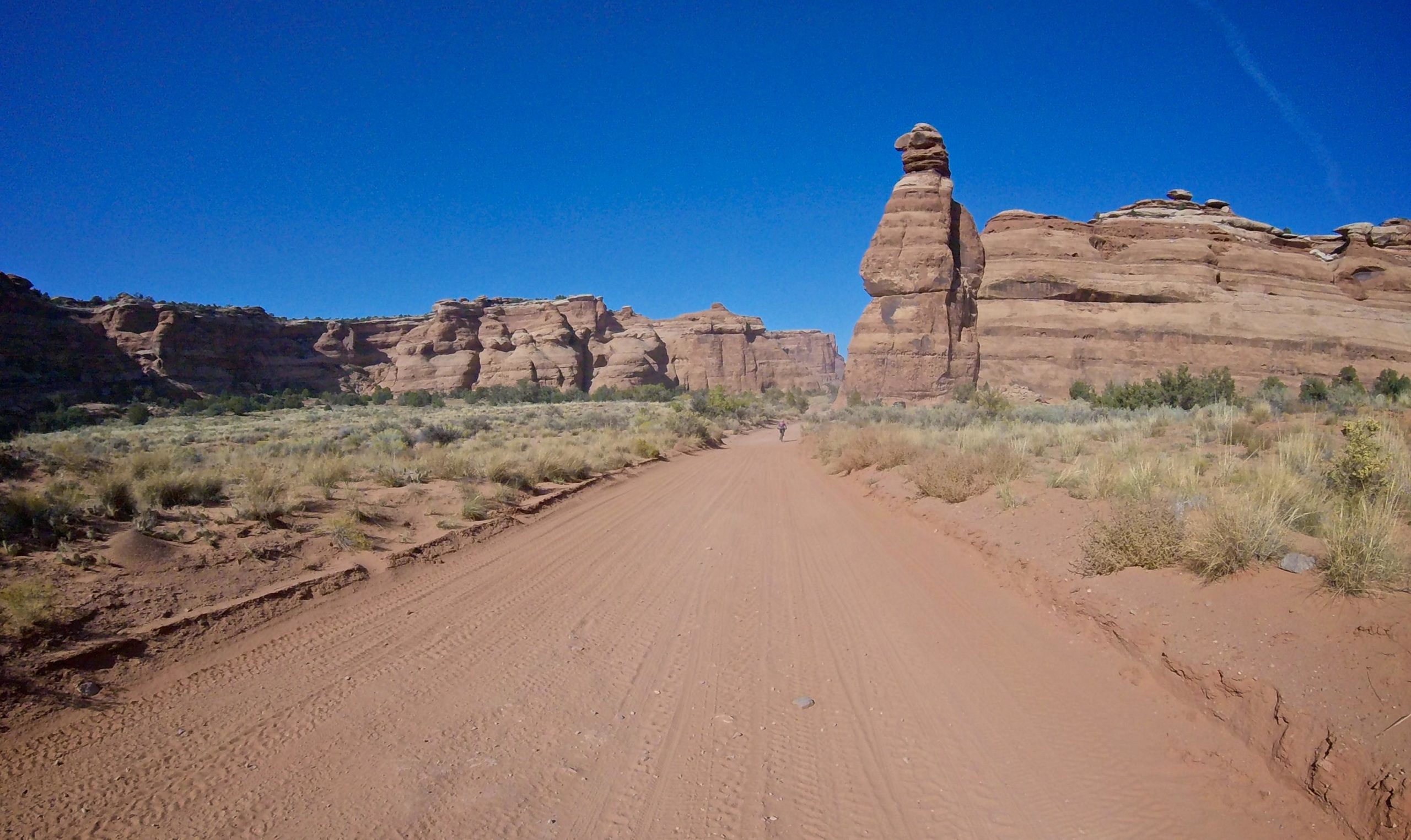 A dirt road winds through a desert landscape, flanked by towering rock formations under a clear blue sky. A prominent rock sculpture resembling a figure stands on the right, while rugged cliffs rise on the left. Sparse vegetation, including patches of grass and shrubs, decorates the sandy terrain.  Gemini Bridges mountain bike trail.