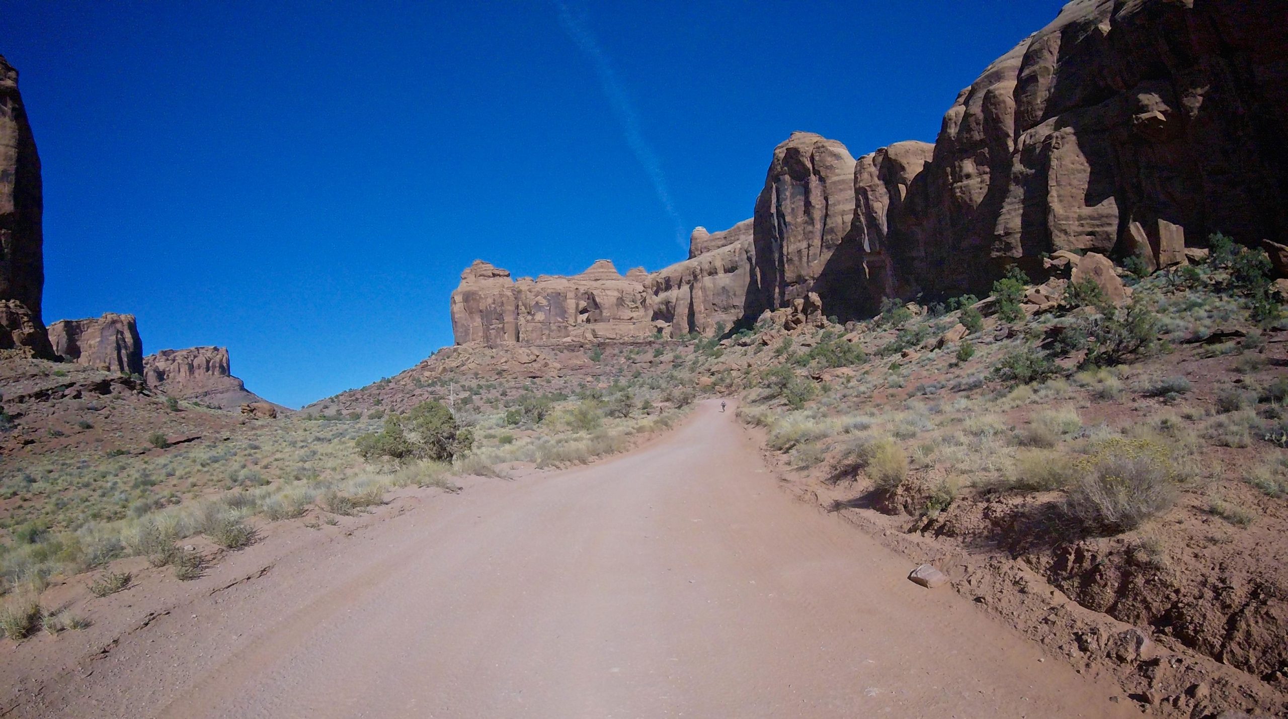A dirt road winding through a rocky landscape under a clear blue sky, surrounded by tall rock formations and sparse vegetation. Gemini Bridges mountain bike trail.