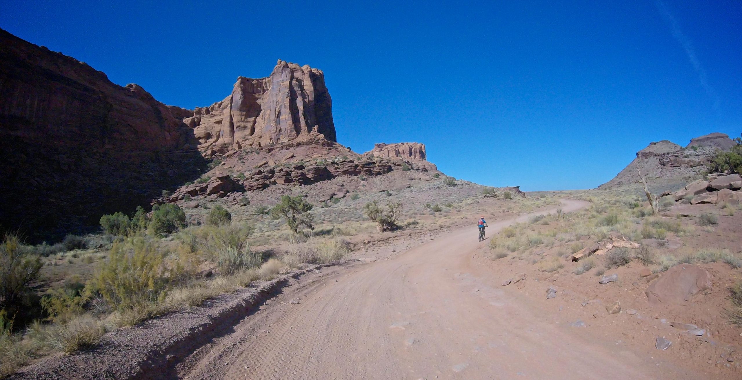 A cyclist riding along a dirt path in a rugged landscape, surrounded by towering rock formations and clear blue skies. Sparse vegetation, including small bushes and grasses, is visible on either side of the trail. Gemini Bridges mountain bike trail.