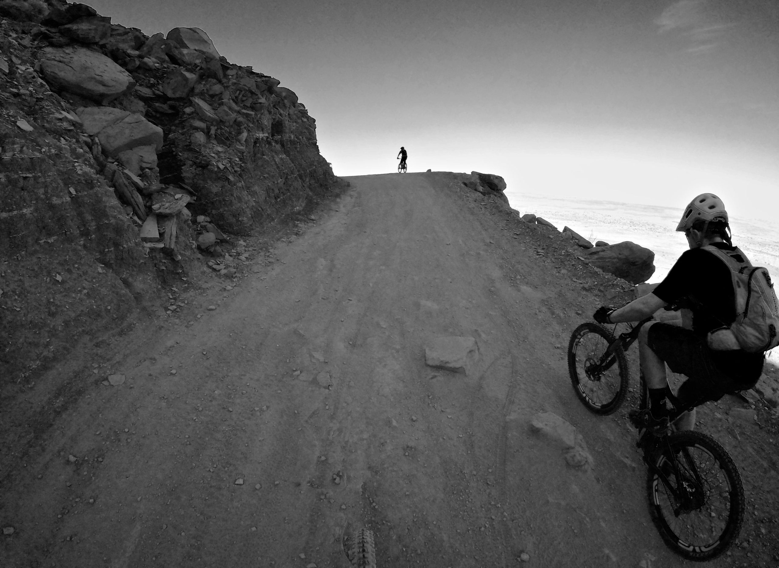 Two mountain bikers riding on a narrow dirt trail surrounded by rocky terrain. One biker is visible in the foreground, slightly leaning forward with a backpack and helmet, while the second biker is further ahead on the trail. The image is presented in black and white, highlighting the rugged landscape and the challenging riding conditions. Gemini Bridges mountain bike trail.