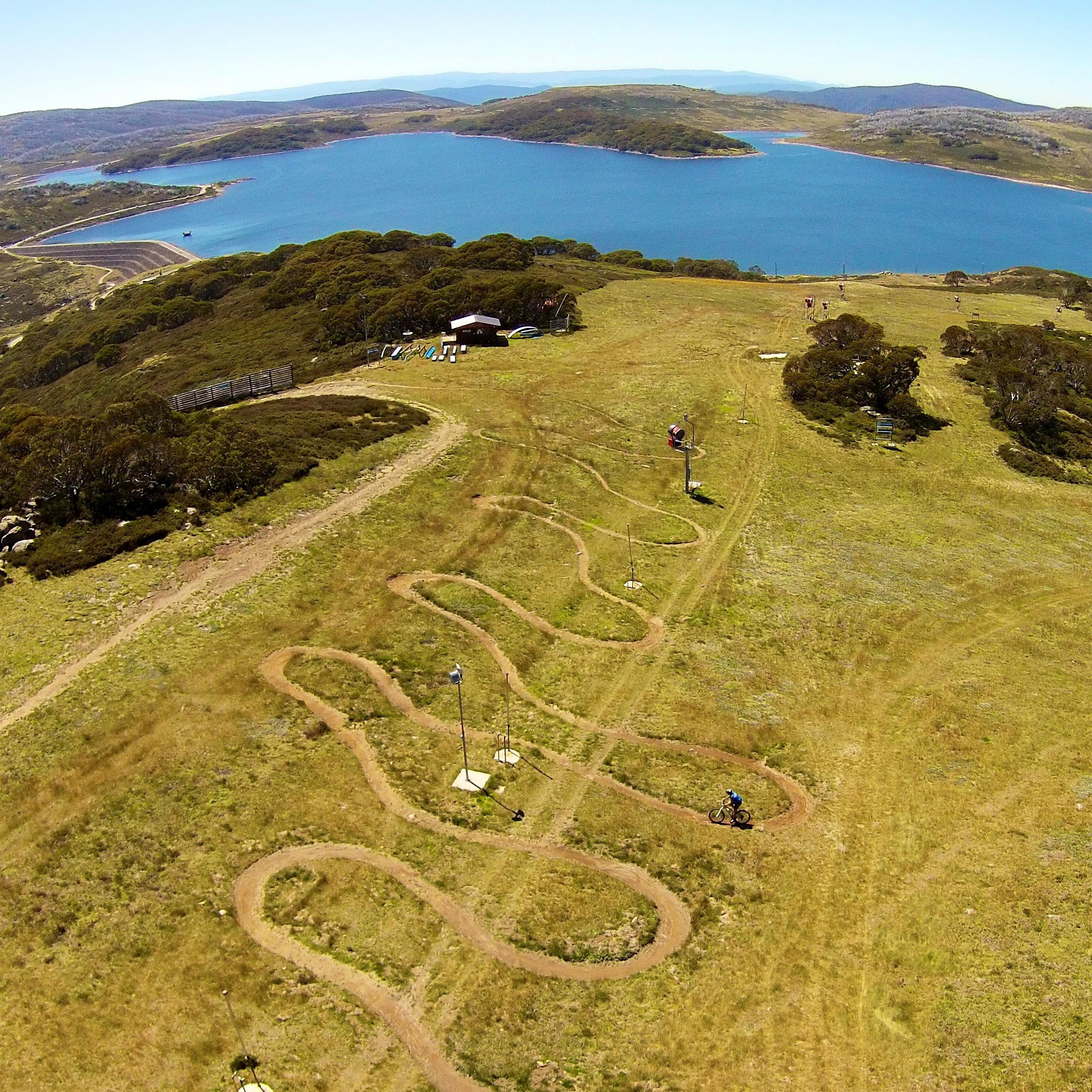 Aerial view of a winding dirt bike track on a grassy hillside, with a blue lake and hills in the background. The track features several loops, and a few small structures are visible nearby. Generator mountain bike trail.