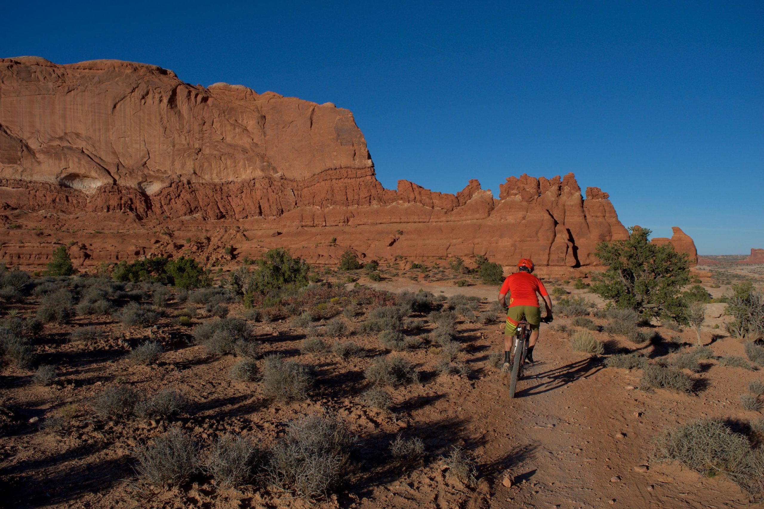 A mountain biker rides along a dirt trail surrounded by red rock formations and sparse vegetation under a clear blue sky. Big Mesa mountain bike trail.