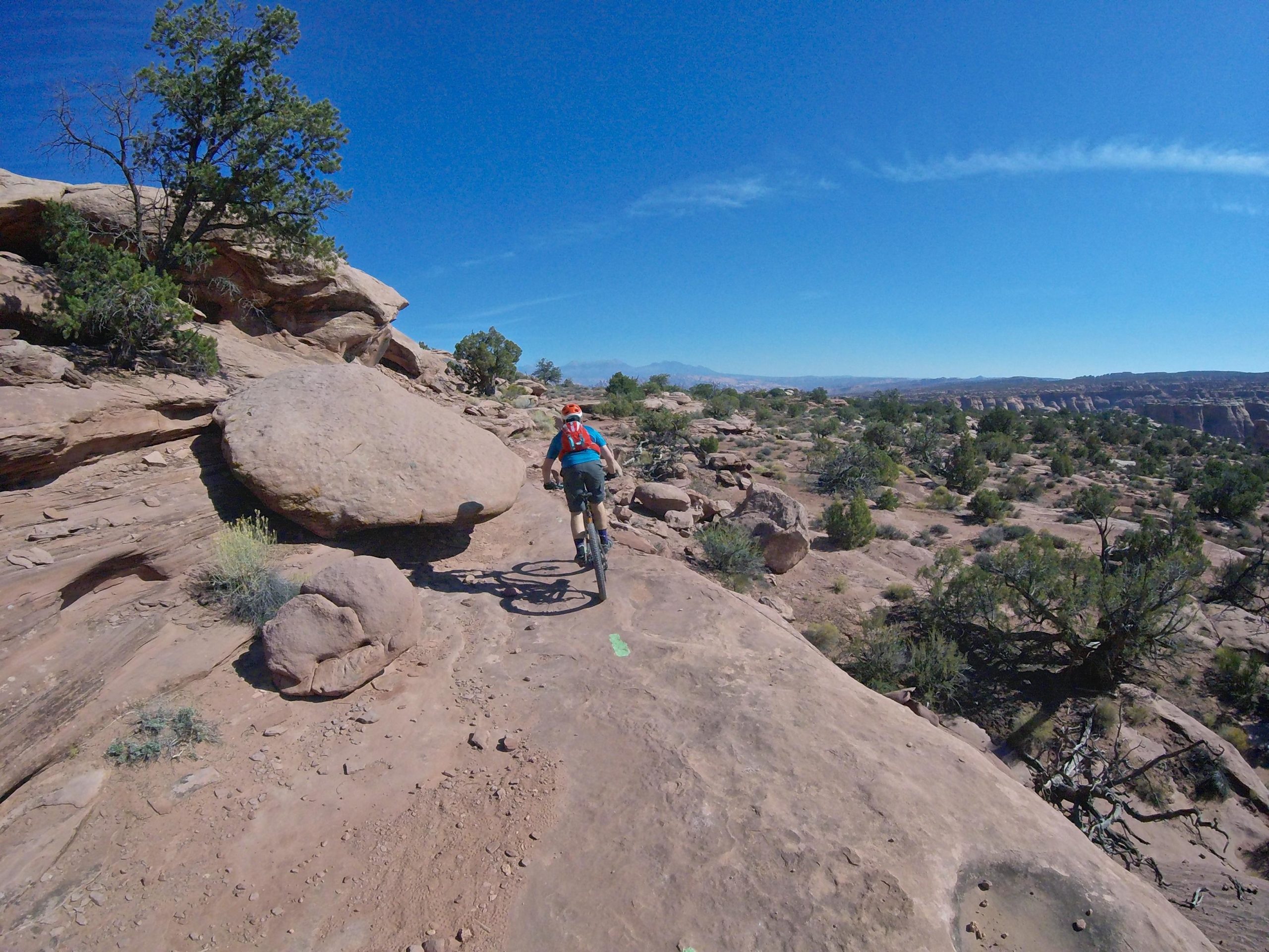 A cyclist riding a mountain bike on a rocky trail surrounded by desert landscape, with large boulders and sparse vegetation under a clear blue sky. Getaway mountain bike trail.