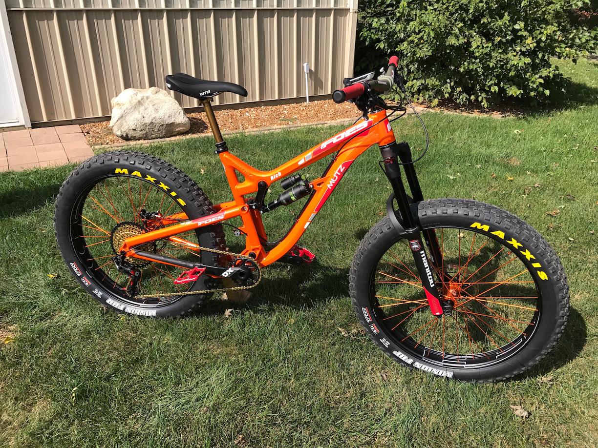 Foes Mutz: A bright orange fat bike parked on green grass, featuring wide tires and an angular frame design. The bike includes red accents on the handlebars and wheels, and the background shows a light-colored building and a large rock.