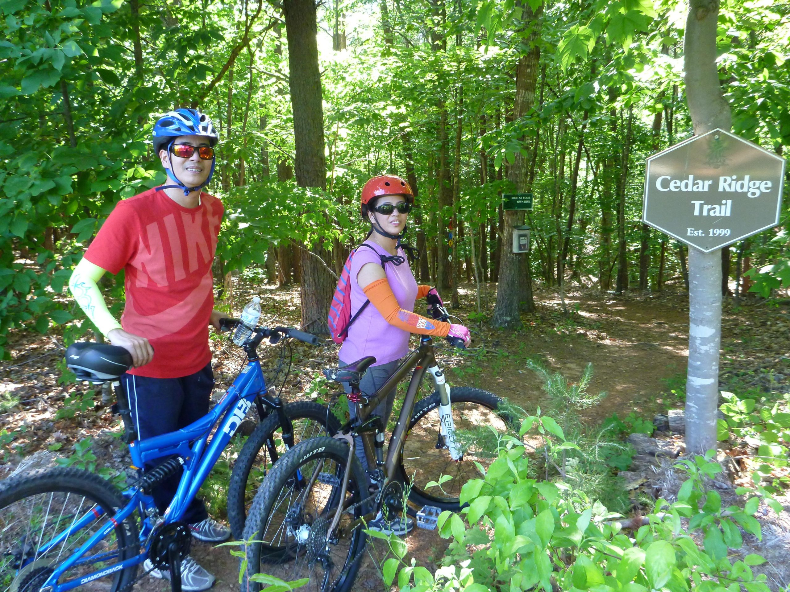 Two cyclists stand next to their mountain bikes at the entrance of Cedar Ridge Trail, surrounded by lush green foliage. The man wears a red t-shirt and a blue helmet, while the woman is dressed in a pink top and an orange long-sleeve shirt, wearing a red helmet and sunglasses. A signpost indicates the name of the trail, established in 1999. Cedar Ridge Chatmoss mountain bike trail.