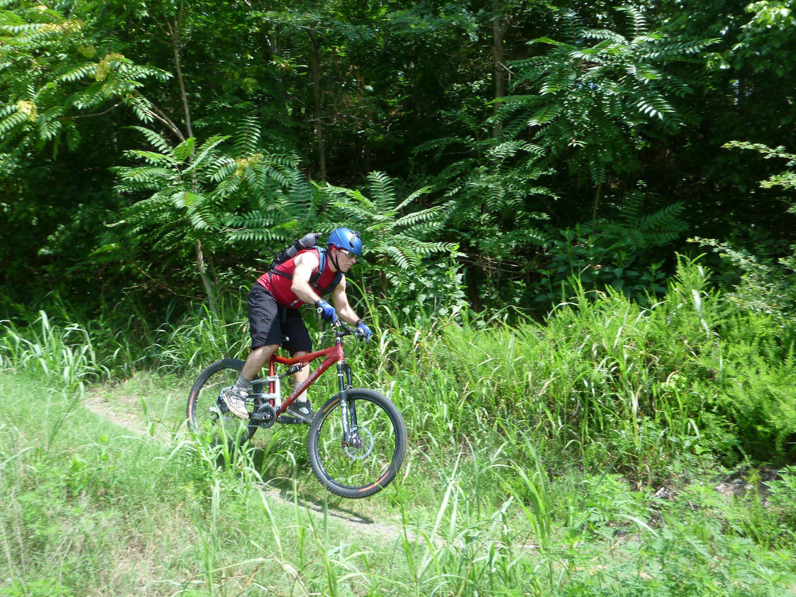 A person wearing a blue helmet and red tank top rides a mountain bike along a dirt trail in a lush green forest. The cyclist is in motion, navigating through tall grass and foliage on either side of the path. Angler's Ridge mountain bike trail.