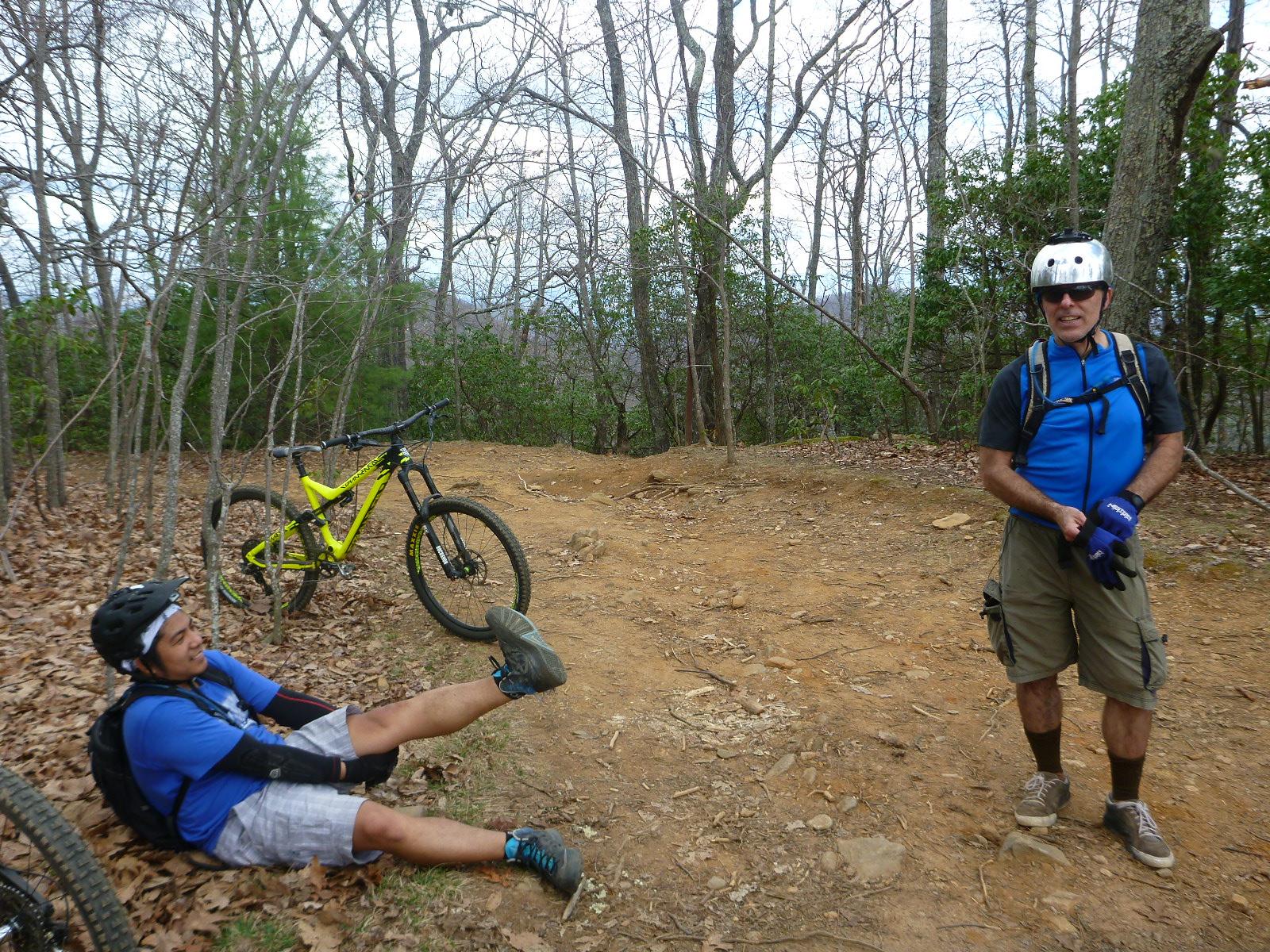 Two mountain bikers are on a dirt trail in a wooded area. One biker, wearing a helmet and blue shirt, is sitting on the ground with his legs raised, appearing to have fallen. The second biker, standing nearby, is wearing a helmet and blue shirt with brown shorts, looking amused or concerned. A bright yellow mountain bike leans against a tree in the background, surrounded by bare trees and scattered leaves on the ground. Bent Creek mountain bike trail.