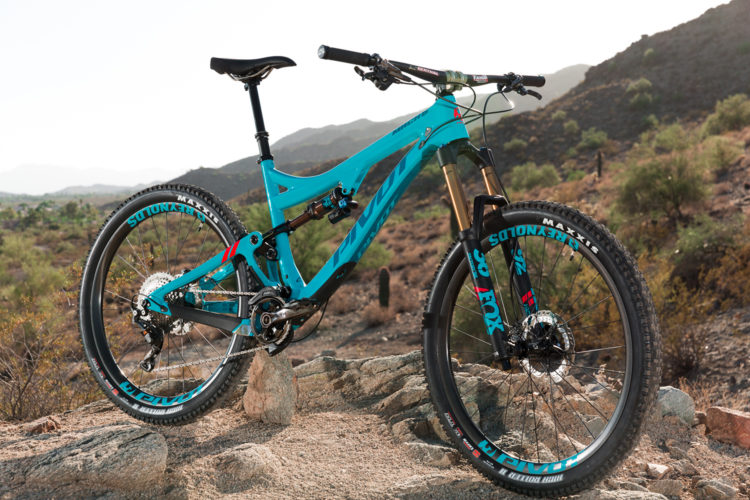 A turquoise mountain bike is positioned on rocky terrain, with a backdrop of hills and sparse vegetation. The bike features a dual suspension system, prominent wheels with branded tires, and a sleek frame design. The scene is illuminated by natural light, emphasizing the bike's construction and detail.