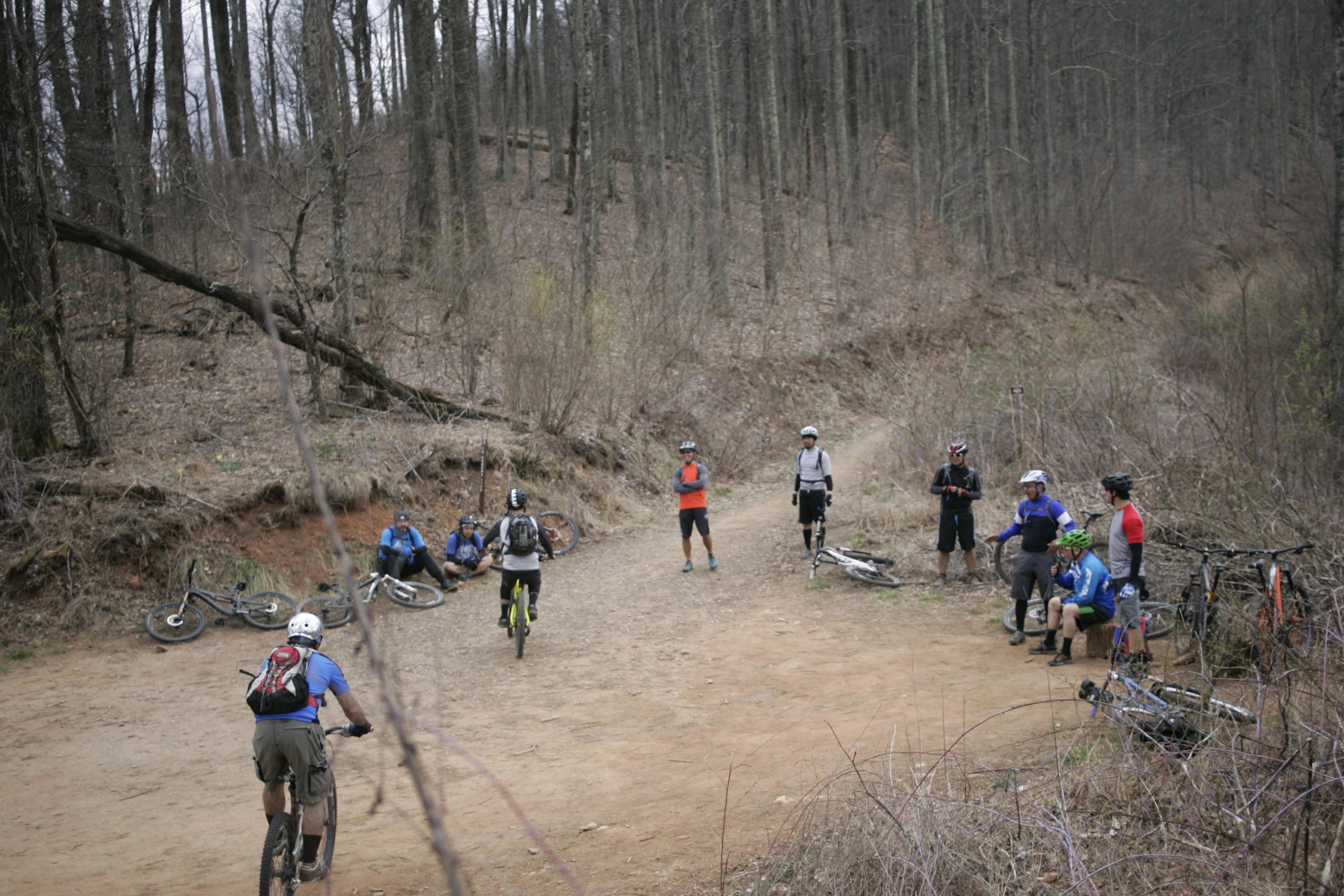 A group of mountain bikers is gathered on a dirt trail surrounded by bare trees, some resting on the ground, while others are standing nearby. Several bicycles are positioned along the trail. The setting appears to be a wooded area, indicative of a mountain biking trail. Bent Creek mountain bike trail.