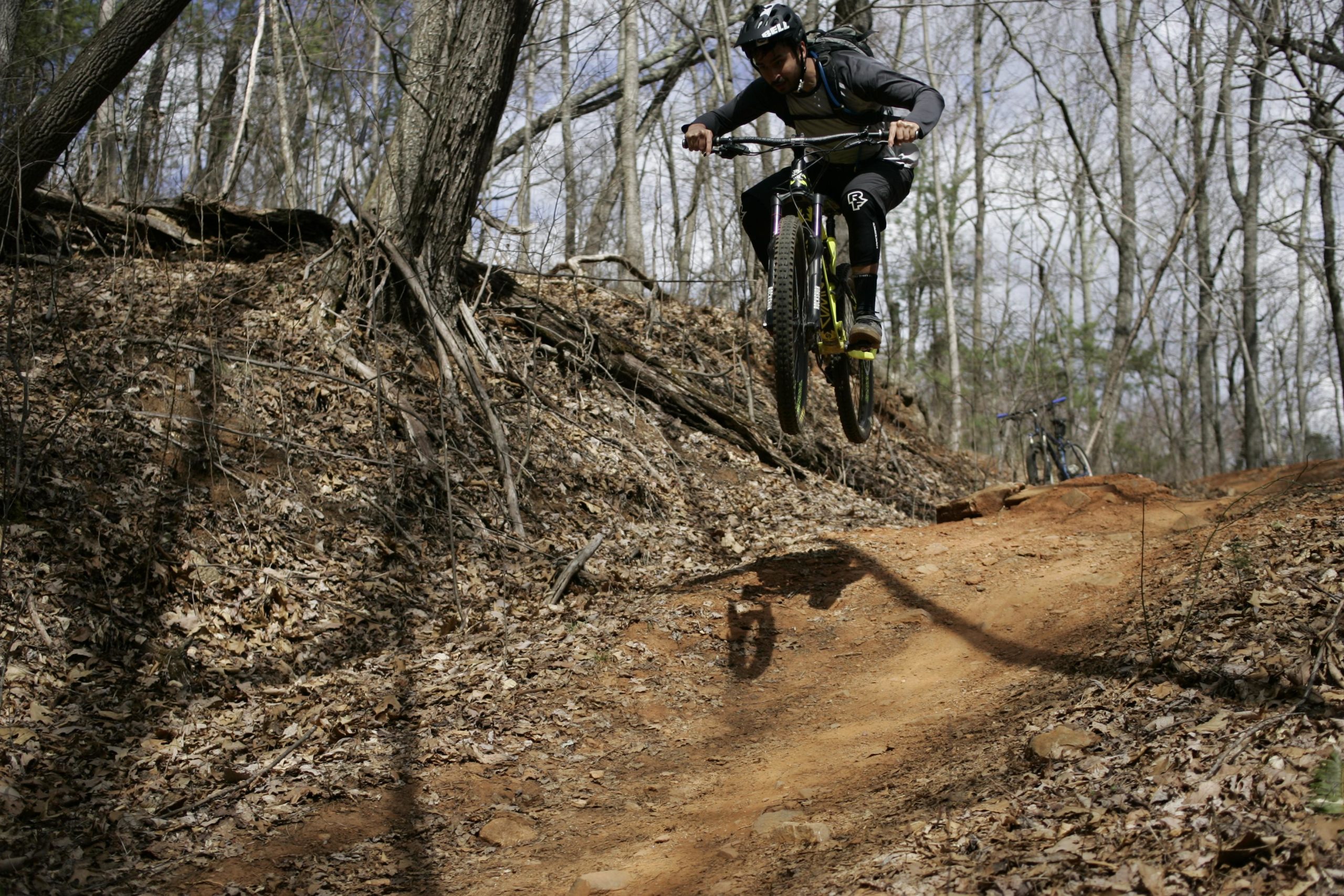 A mountain biker performs a jump on a dirt trail surrounded by trees, with leaves scattered on the ground. The biker is wearing a helmet and a backpack, demonstrating an adventurous stance mid-air. A second bike is visible in the background. Bent Creek mountain bike trail.