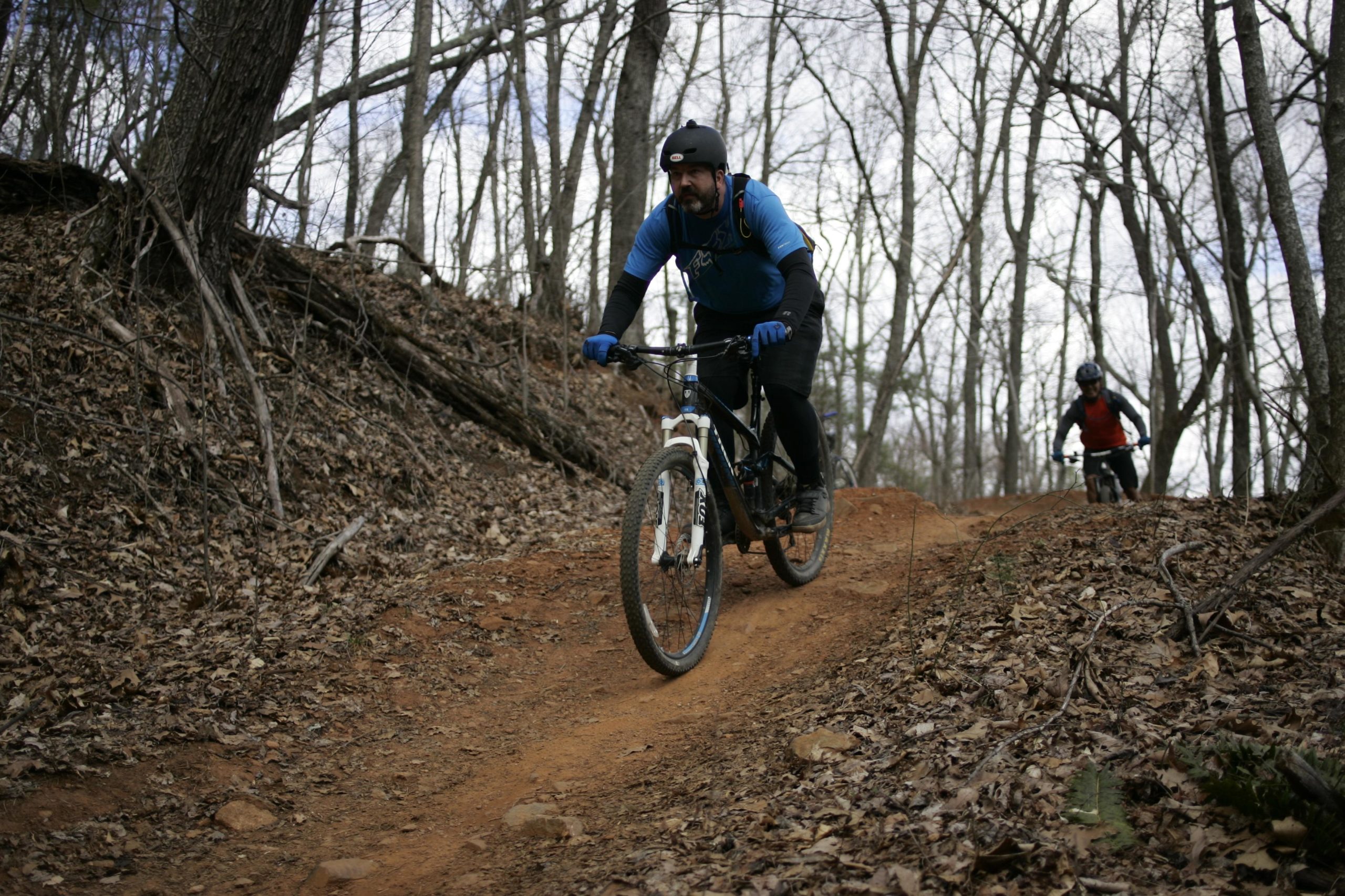 A man in a blue shirt and black shorts rides a mountain bike down a dirt trail surrounded by bare trees and fallen leaves. Another cyclist can be seen in the background, navigating the same path. The scene captures an outdoor biking adventure in a natural environment. Bent Creek mountain bike trail.