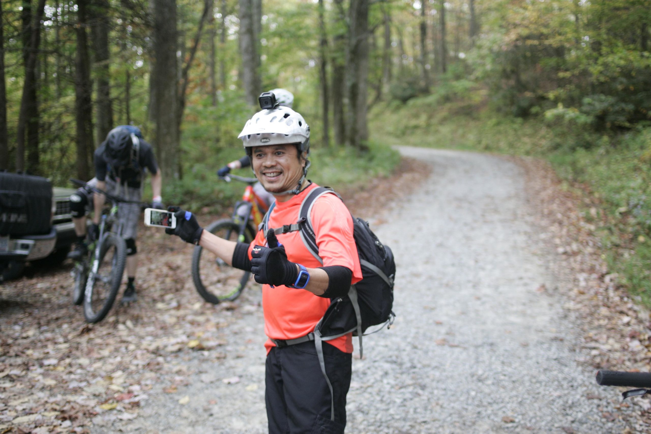 A smiling cyclist in an orange shirt and helmet stands on a gravel path in a wooded area, giving a thumbs-up while holding a smartphone. In the background, another cyclist is adjusting their bike, surrounded by autumn foliage. Bennett Gap / 138 mountain bike trail.