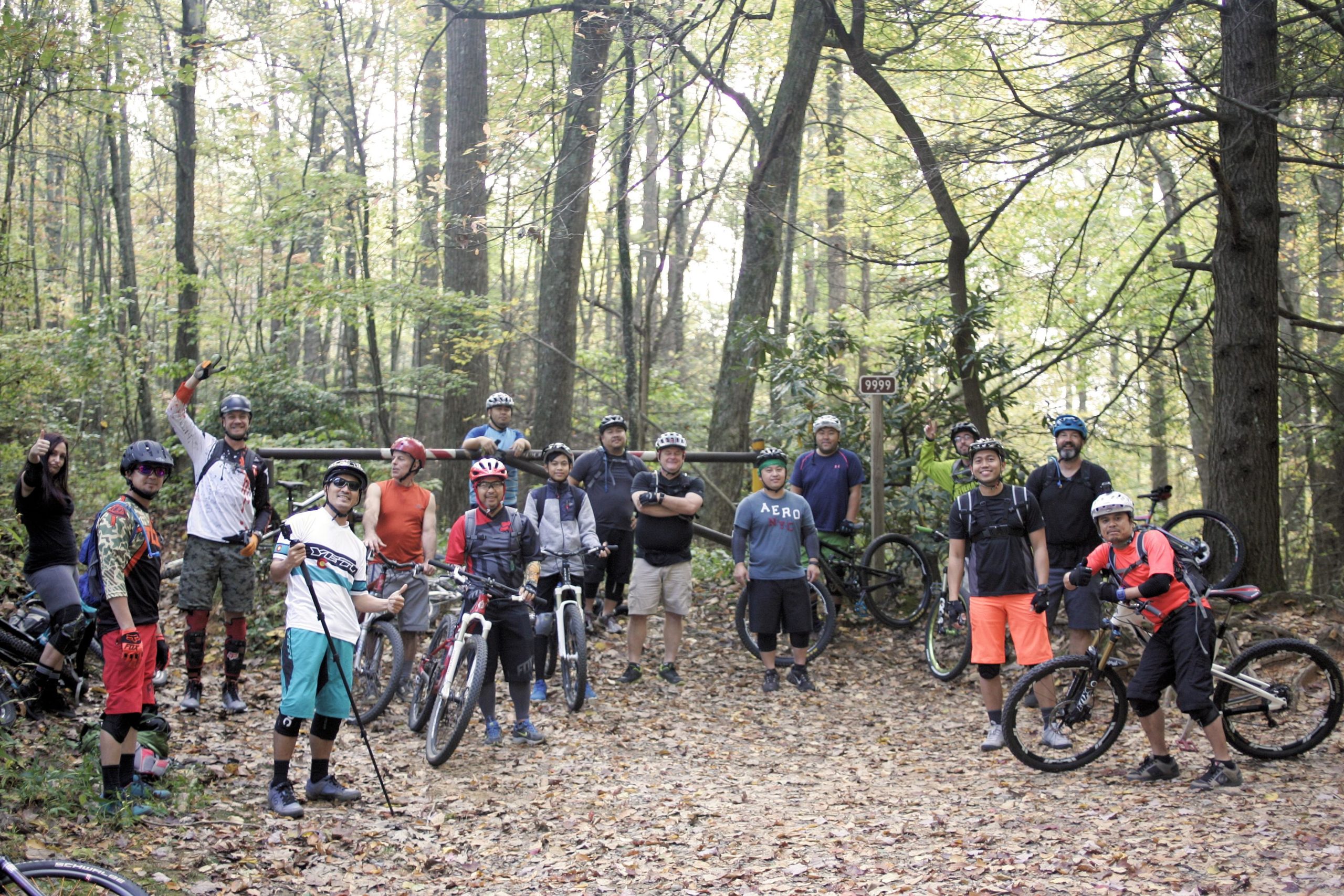 A group of mountain bikers poses together on a trail surrounded by autumn foliage. Some are giving thumbs up or smiling at the camera. They are wearing biking gear and helmets, with their bicycles parked next to them. A trail sign labeled "9999" is visible in the background, indicating the path they are on. Bennett Gap / 138 mountain bike trail.