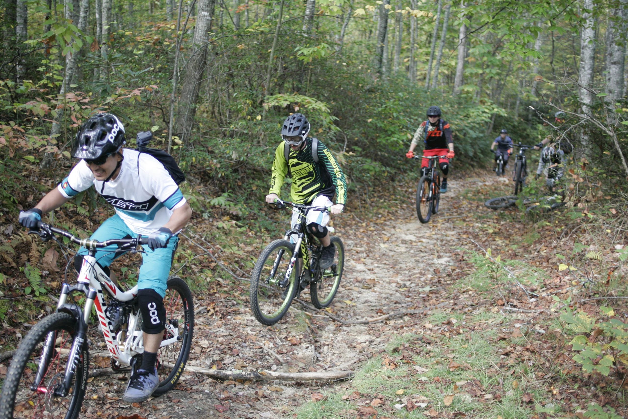 A group of four mountain bikers navigating a dirt trail surrounded by trees and autumn foliage. Two riders are in the foreground, one wearing a white and teal jersey and another in a green and black jersey. The other two riders are further back, with visible bikes at various points along the trail. The terrain is uneven, with patches of grass and fallen leaves. Bennett Gap / 138 mountain bike trail.