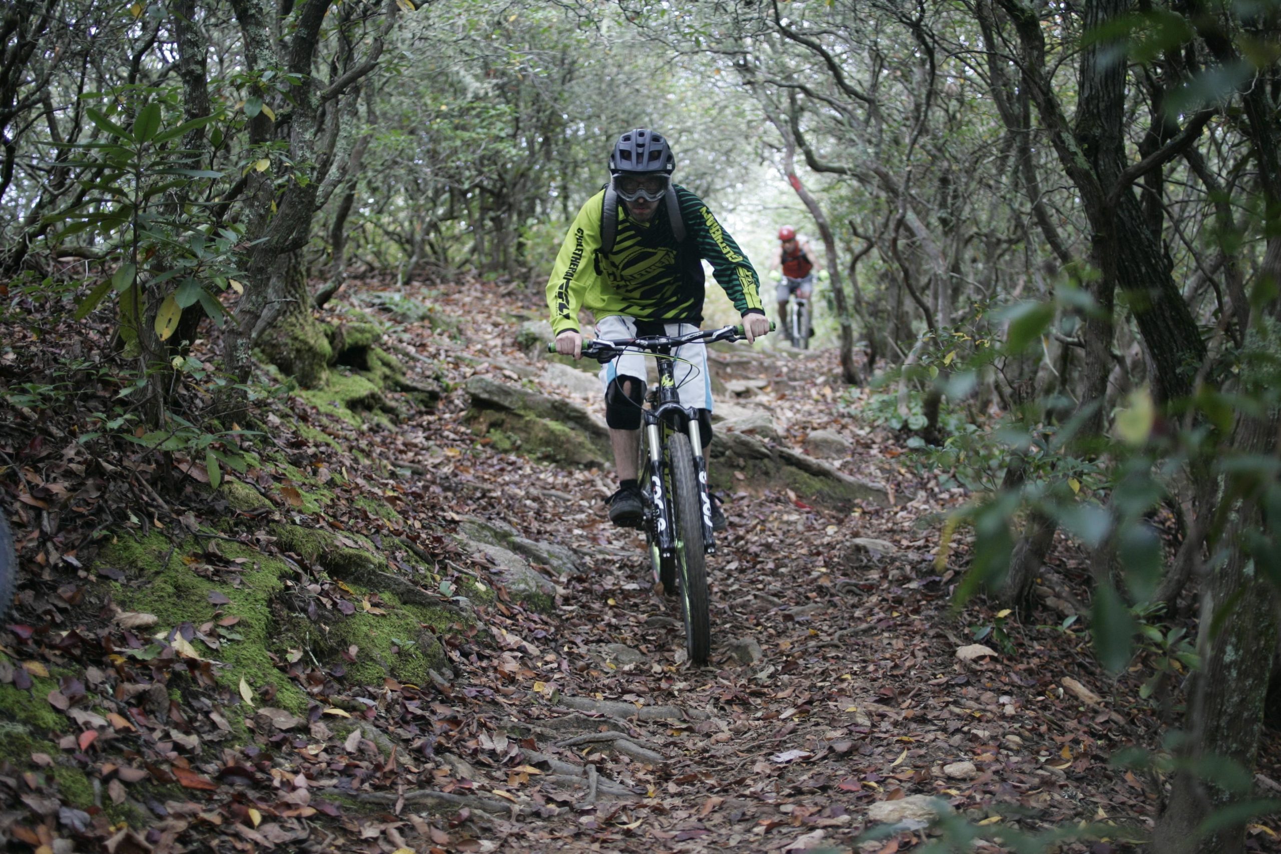 A mountain biker rides along a narrow, rocky trail surrounded by dense foliage and fallen leaves. One bike is in the foreground, with the rider wearing a bright green long-sleeve jersey and a helmet. Another biker is seen further back on the same trail. The scene captures the essence of outdoor adventure in a natural setting. Bennett Gap / 138 mountain bike trail.