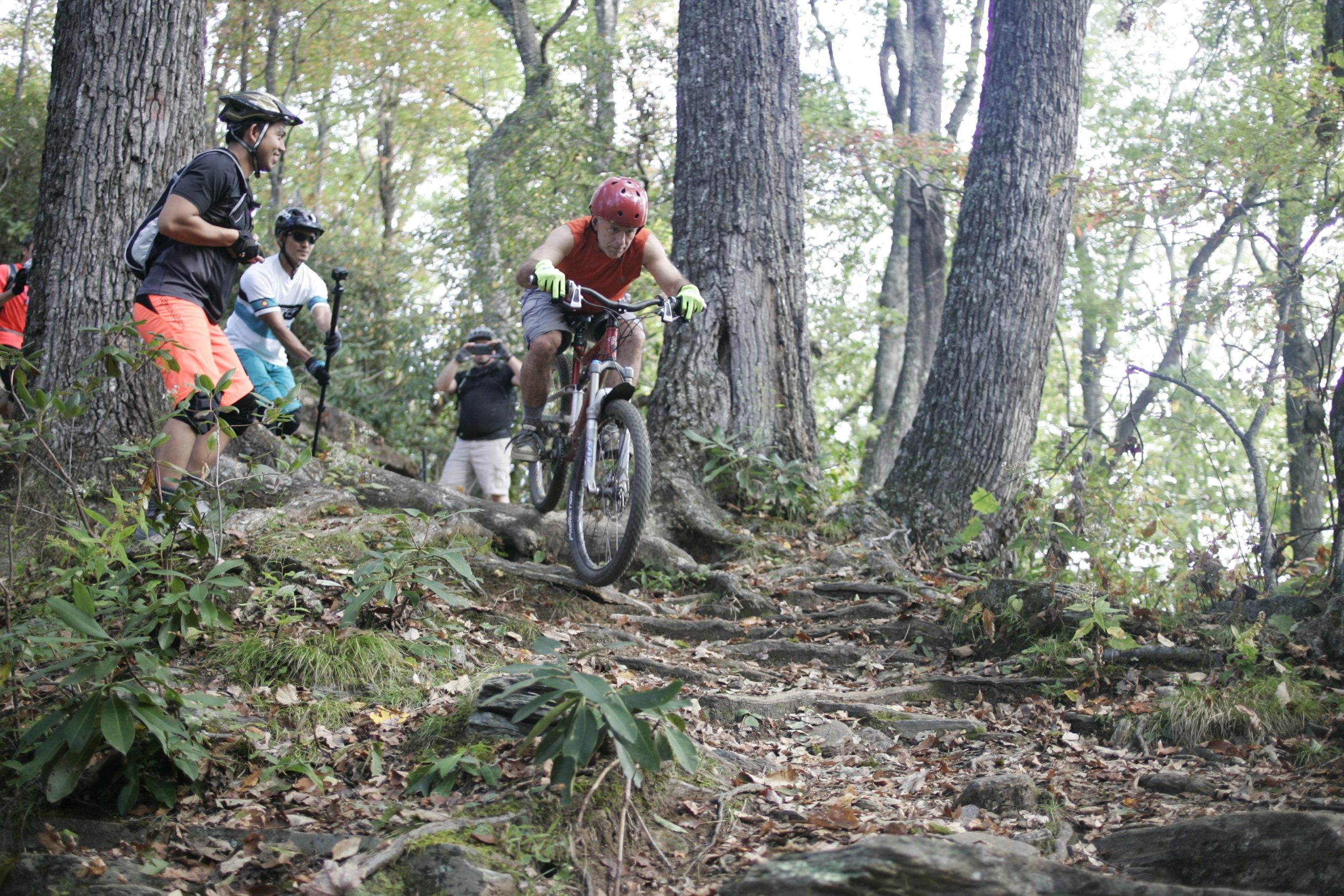 A mountain biker in a red helmet and sleeveless red shirt navigates over rocky terrain in a forested area, while several onlookers watch and cheer from the side. The scene captures the excitement of outdoor biking on a trail surrounded by tall trees and greenery. Bennett Gap / 138 mountain bike trail.