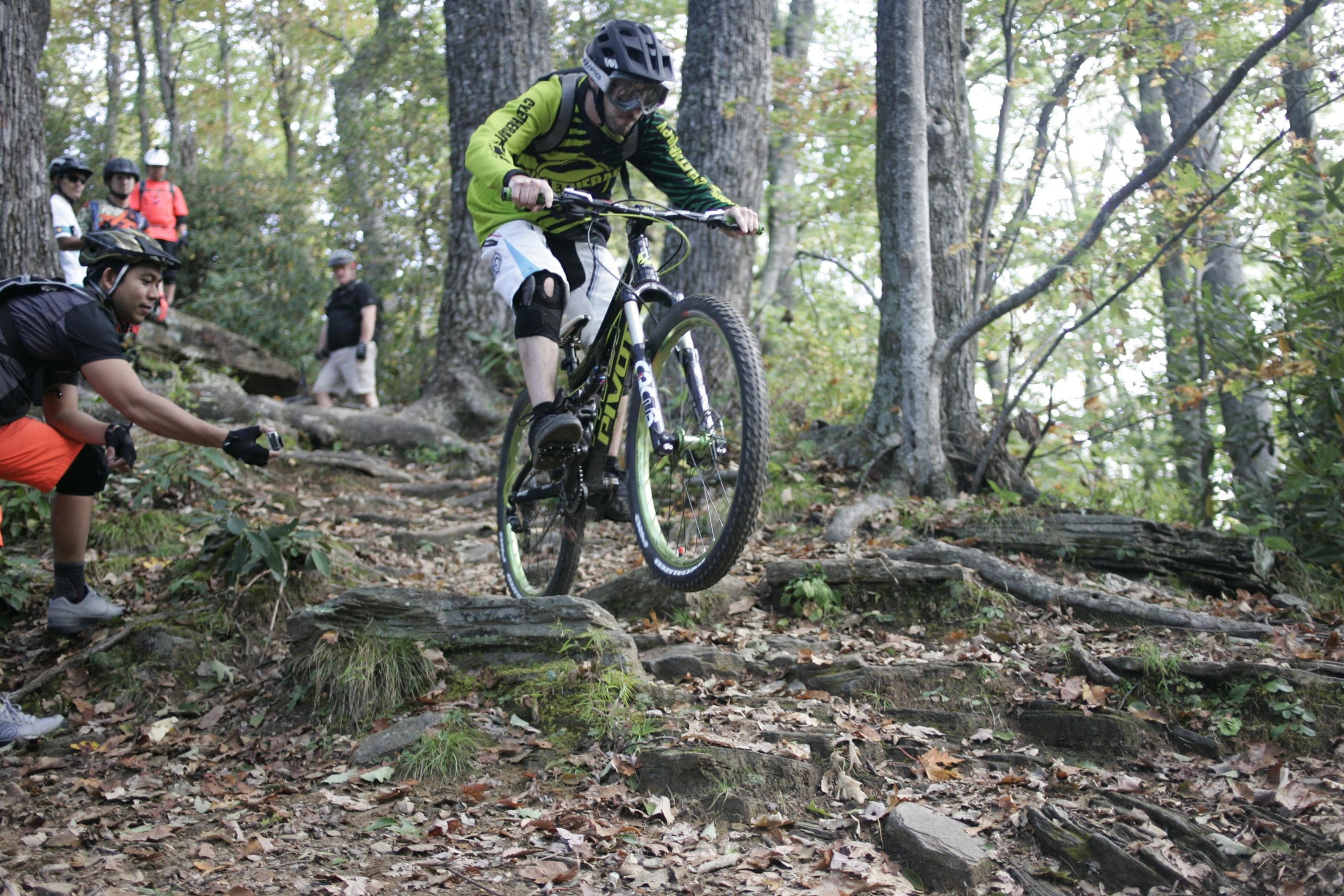 A mountain biker in a green jersey and protective gear maneuvers over rocky terrain while performing a jump. A man in a black shirt and orange shorts reaches out with a camera, capturing the action, while other spectators watch in the background amidst the trees. Bennett Gap / 138 mountain bike trail.