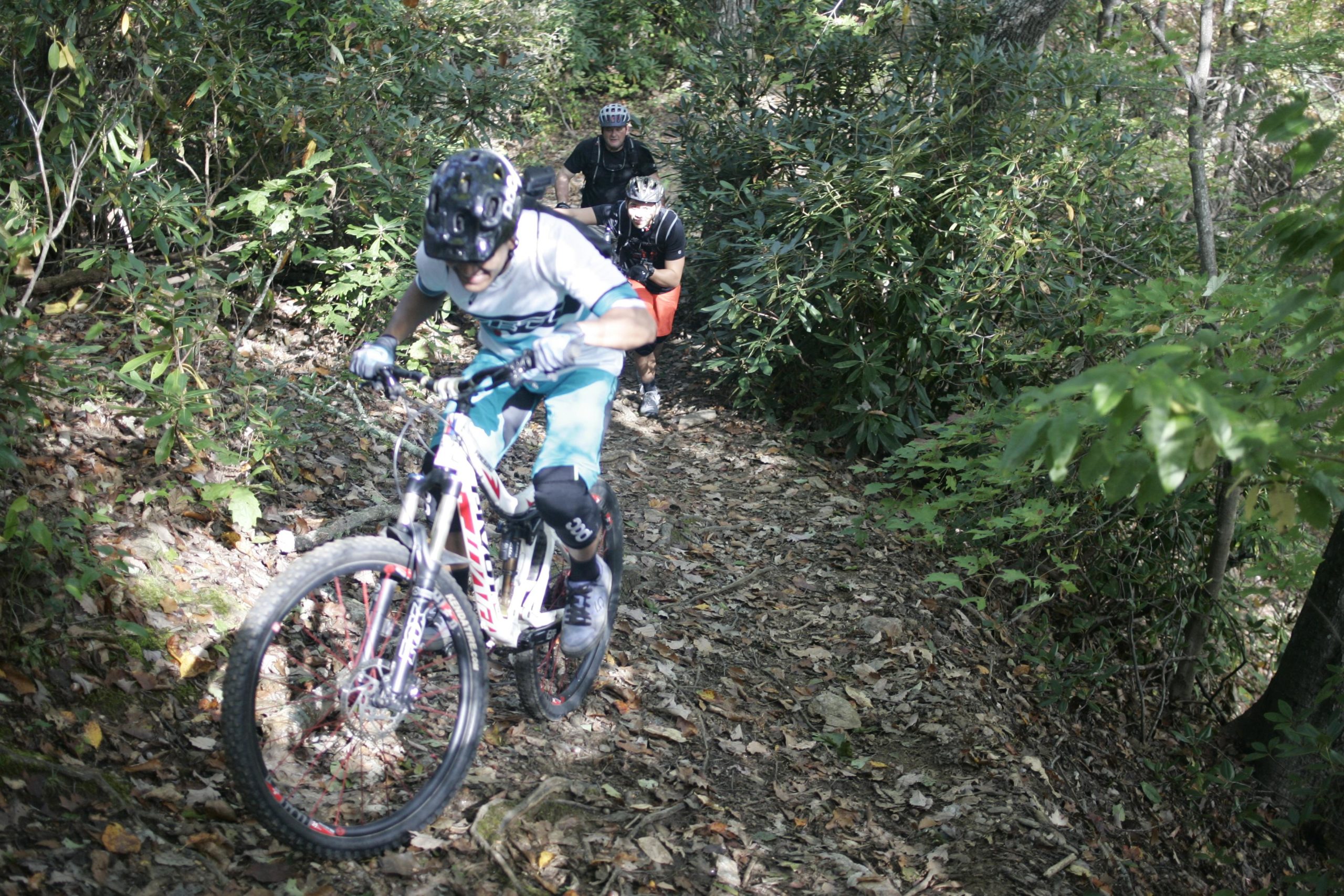 A group of mountain bikers navigating a narrow trail through a wooded area, surrounded by greenery and fallen leaves. The lead cyclist is actively riding, while two others follow closely behind, ensuring they stay on the path. Bennett Gap / 138 mountain bike trail.
