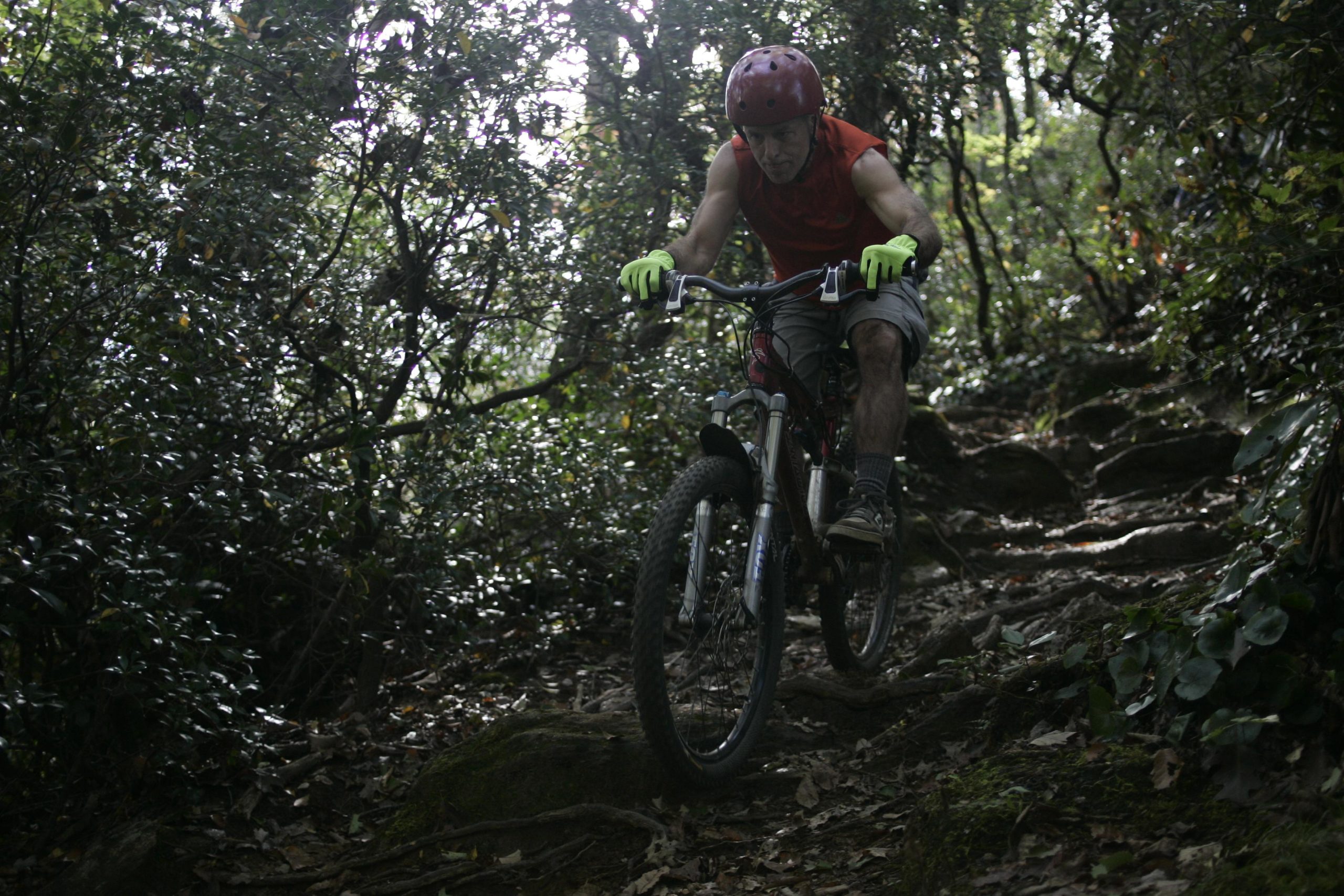 A mountain biker navigating a rocky trail in a dense, wooded area, wearing a bright red helmet, green gloves, and a sleeveless red shirt. Bennett Gap / 138 mountain bike trail.
