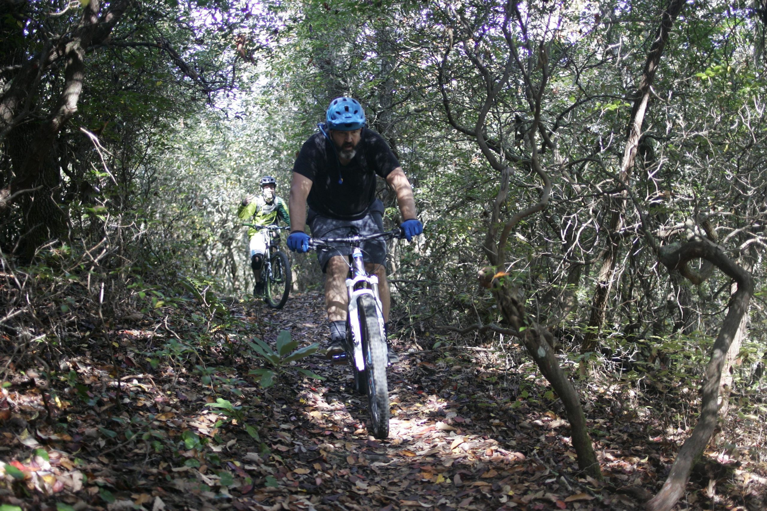 Two mountain bikers navigate a narrow, wooded trail. The first rider, wearing a blue helmet and gloves, is focused on the path ahead. The second rider, dressed in green, follows behind, giving a thumbs up. The ground is covered with fallen leaves, and dense foliage surrounds the trail. Bennett Gap / 138 mountain bike trail.