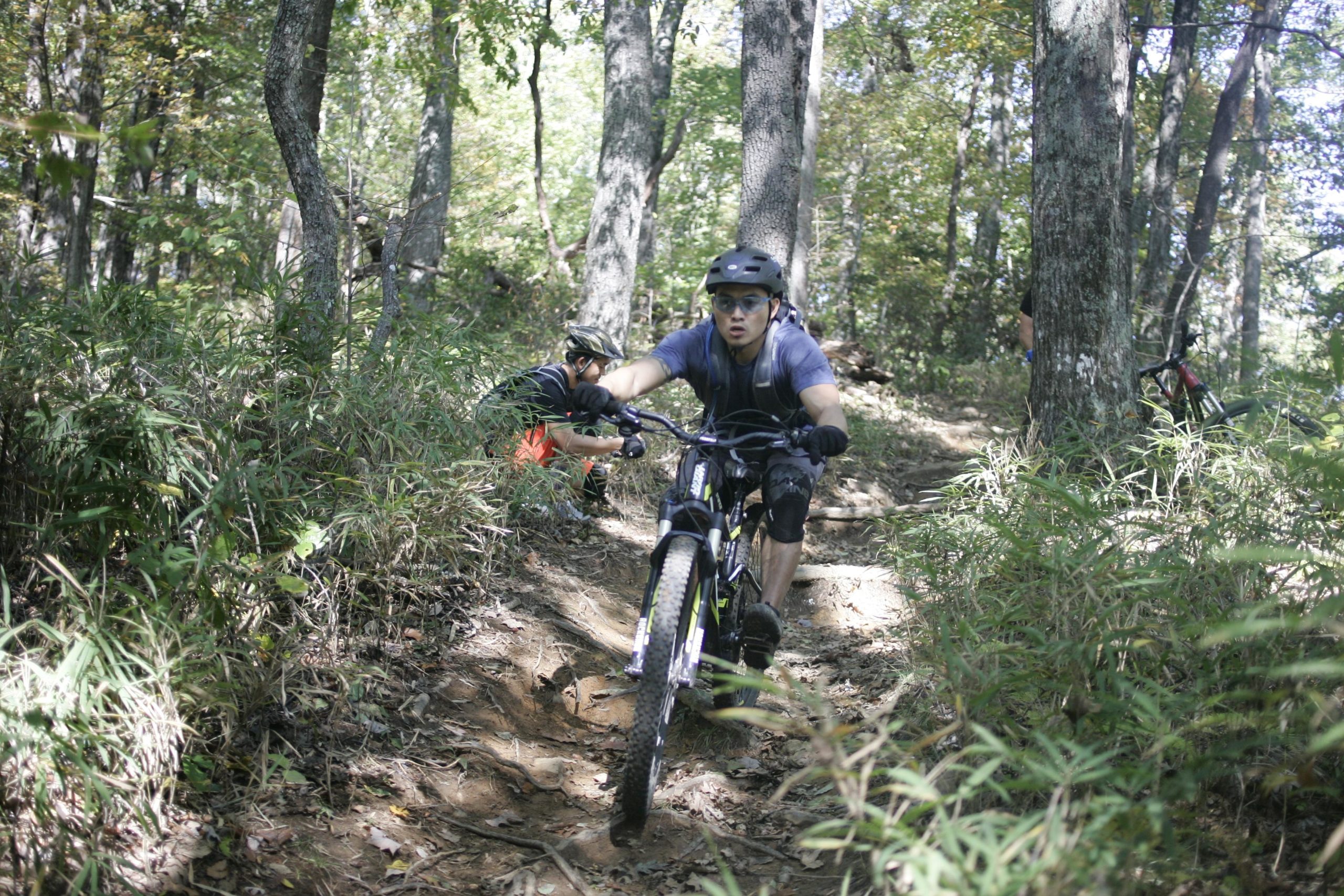 A mountain biker rides along a dirt trail surrounded by trees and greenery, showing focus and determination. In the background, another person is crouched down, possibly adjusting their bike. The scene captures an active outdoor adventure in a wooded area. Bennett Gap / 138 mountain bike trail.