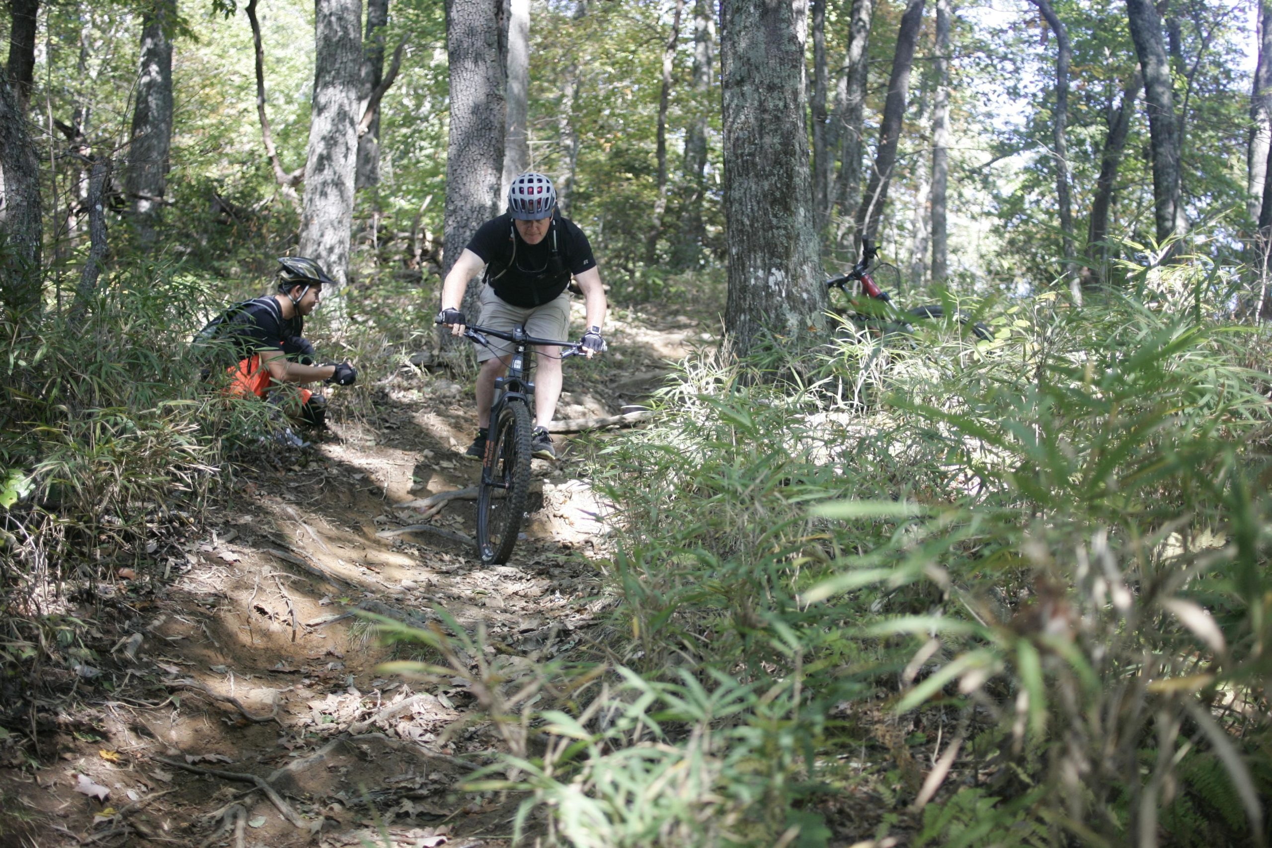 A mountain biker navigates a rugged dirt trail in a wooded area, while a person in the background sits beside the trail with a camera, capturing the action. The scene is surrounded by greenery and trees, showcasing a sunny day in nature. Bennett Gap / 138 mountain bike trail.