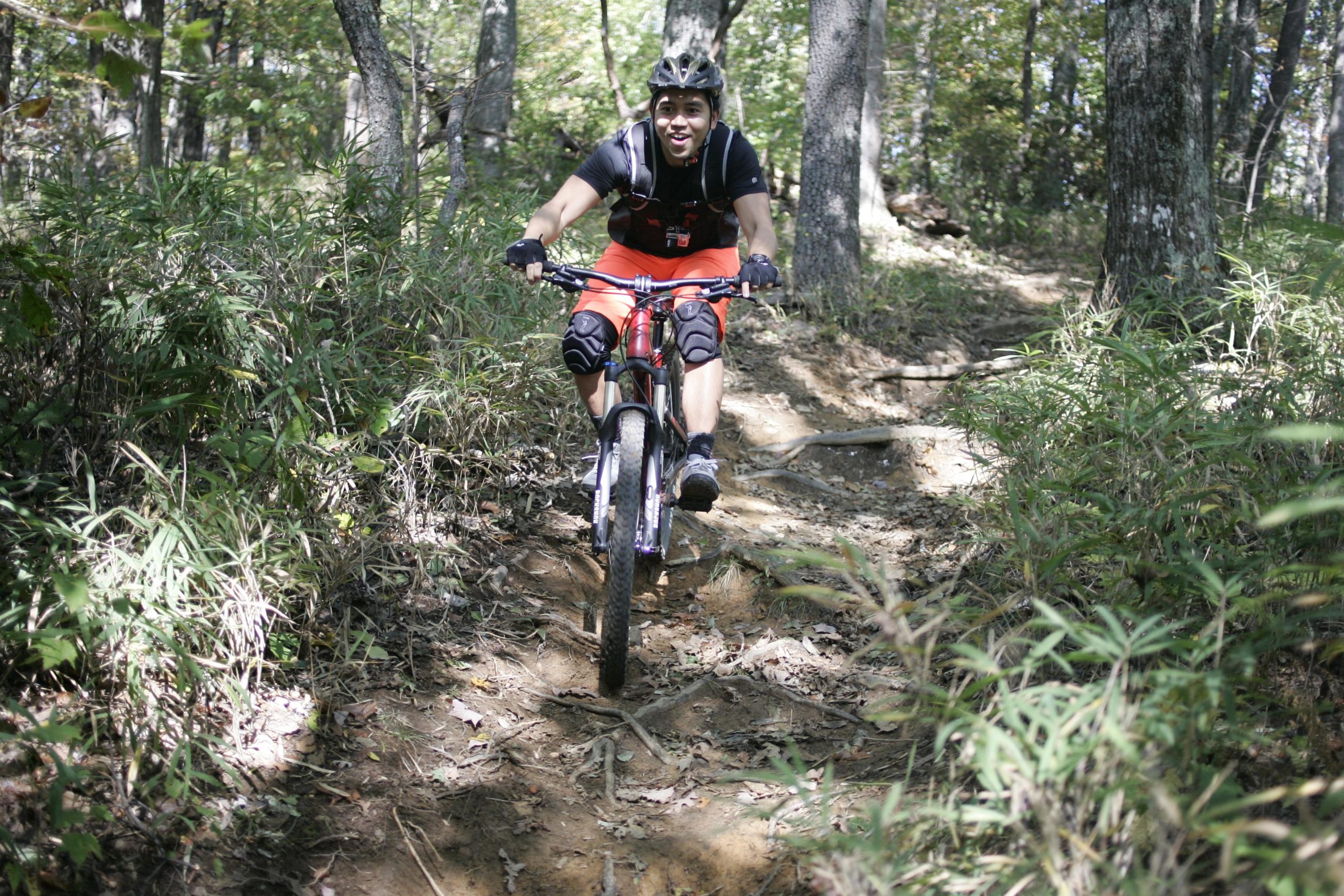 A person riding a mountain bike down a dirt trail surrounded by trees and greenery, wearing a helmet, gloves, and protective gear, with a smile on their face. Bennett Gap / 138 mountain bike trail.