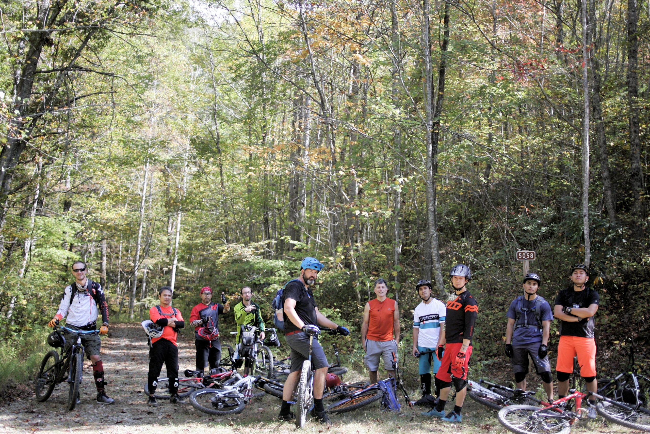 A group of ten mountain bikers gathered on a forest trail, smiling and posing for the camera. They are dressed in various cycling gear, including helmets and gloves, and several bikes are parked nearby. The background features tall trees with green leaves, indicating a sunny day. A trail marker sign is visible, adding a sense of location to the scene. Black Mountain mountain bike trail.