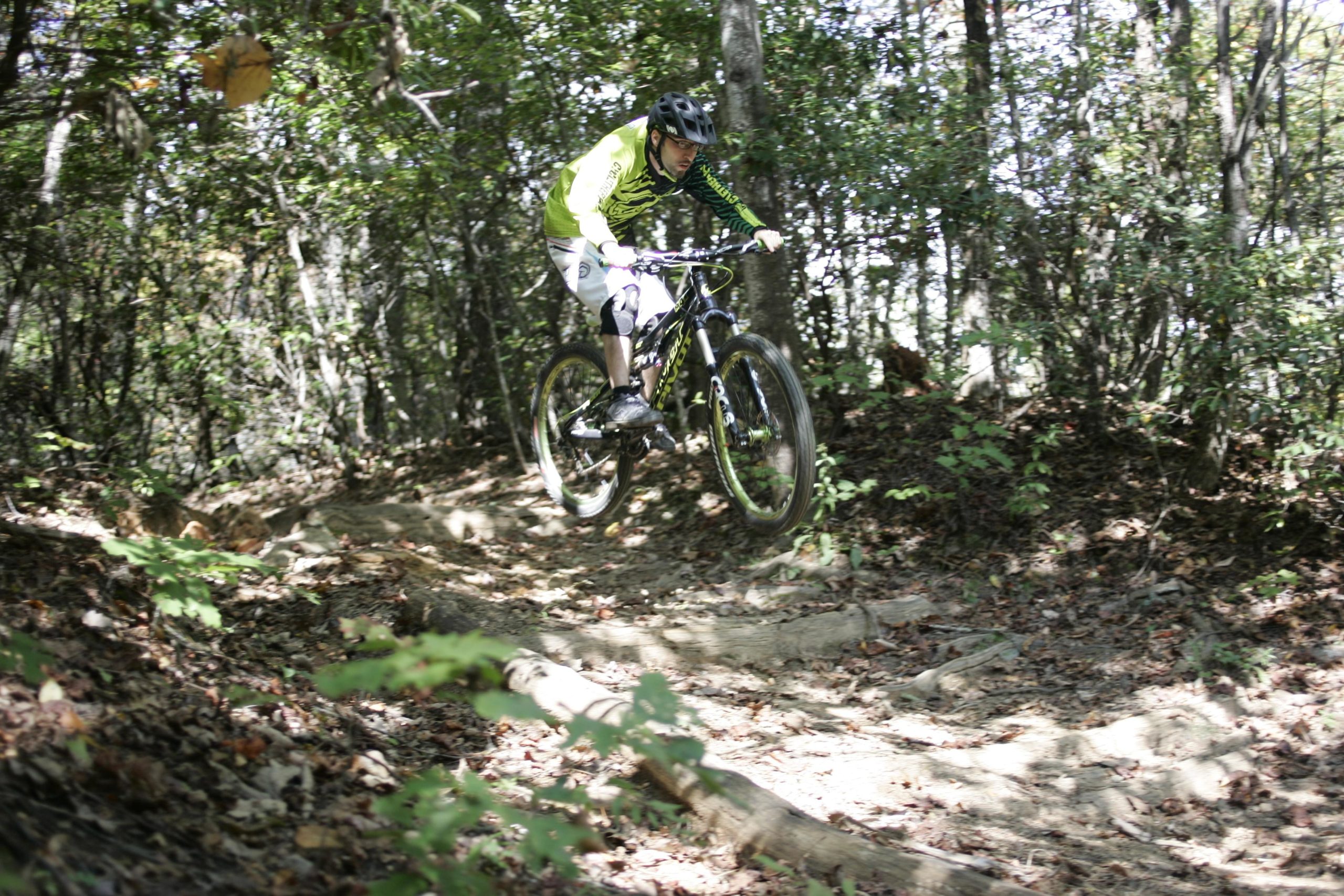 A mountain biker in a bright yellow and black jersey jumps over a rocky trail surrounded by trees in a forested area. Black Mountain mountain bike trail.