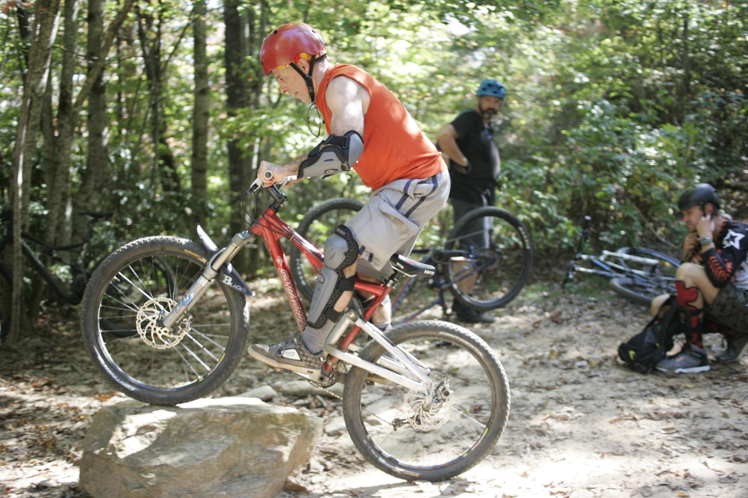 A mountain biker in an orange shirt and red helmet is performing a wheelie by balancing on a rock, while two other cyclists are watching nearby in a forested area. Bennett Gap / 138 mountain bike trail.