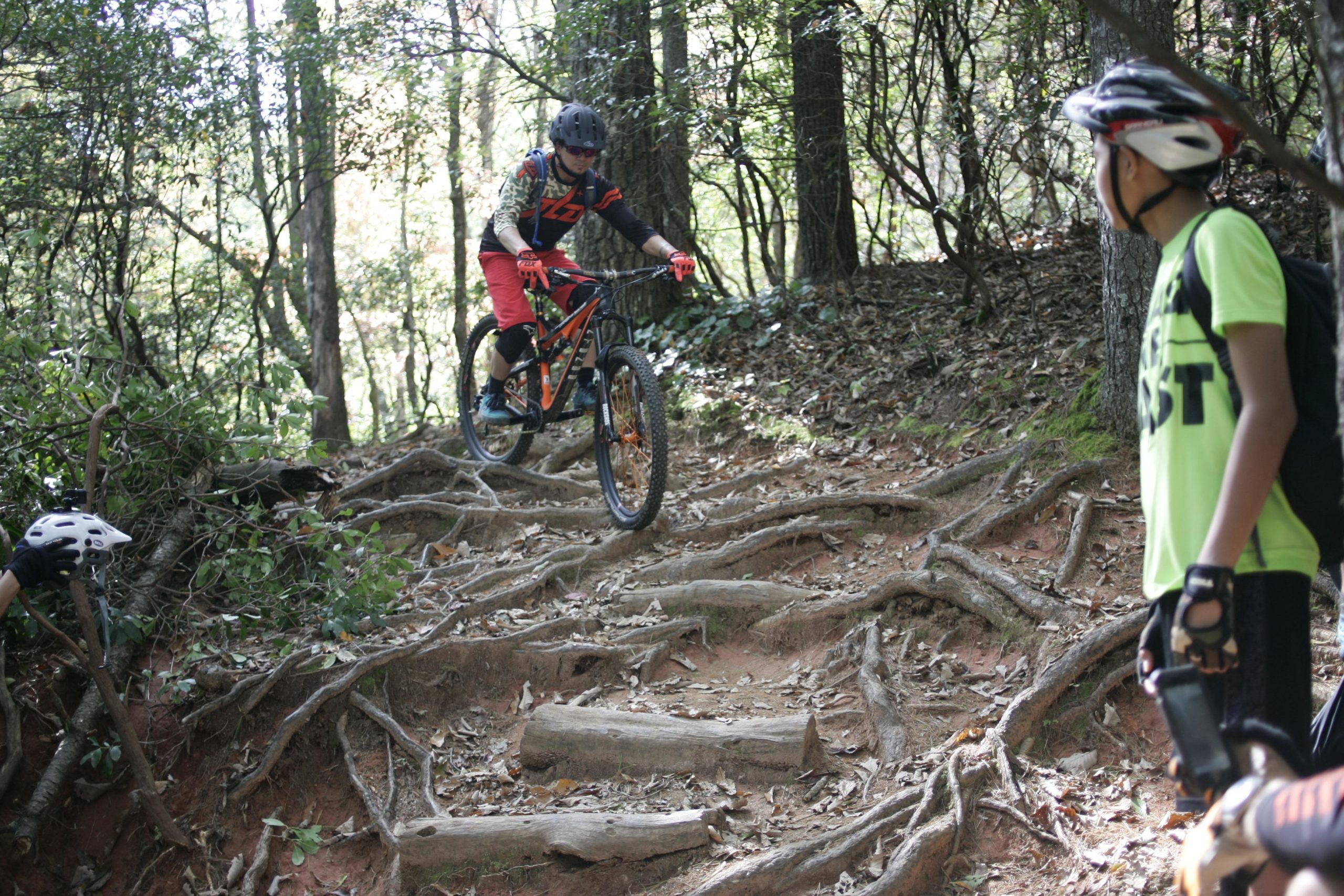 A mountain biker navigating a rocky and root-covered trail in a forest, with two other riders observing nearby. One is wearing a helmet and gloves, while the other is dressed in a bright green shirt and has a backpack. Sunlight filters through the trees, highlighting the natural surroundings. Bennett Gap / 138 mountain bike trail.