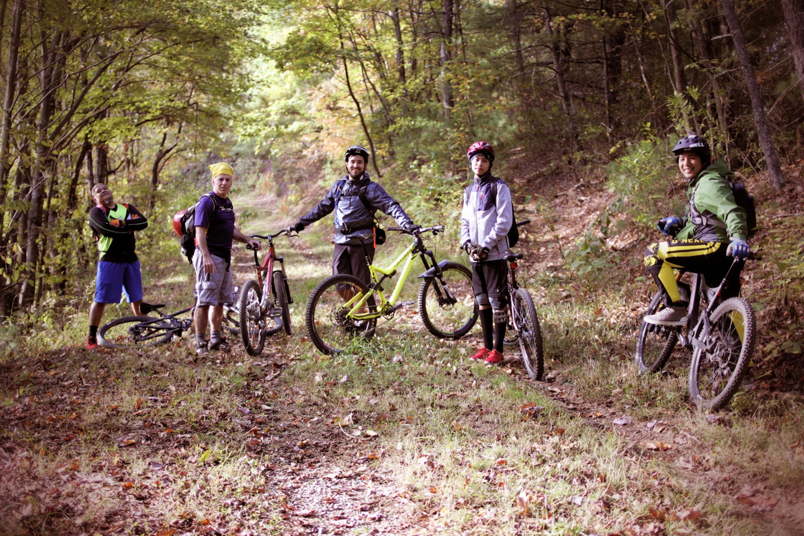 A group of five mountain bikers standing on a dirt path in a forested area, surrounded by trees with autumn foliage. They are posing with their bicycles, wearing cycling gear, and appear to be enjoying a break in their ride. The scene captures the camaraderie of outdoor biking in nature. Sinkhole mountain bike trail.