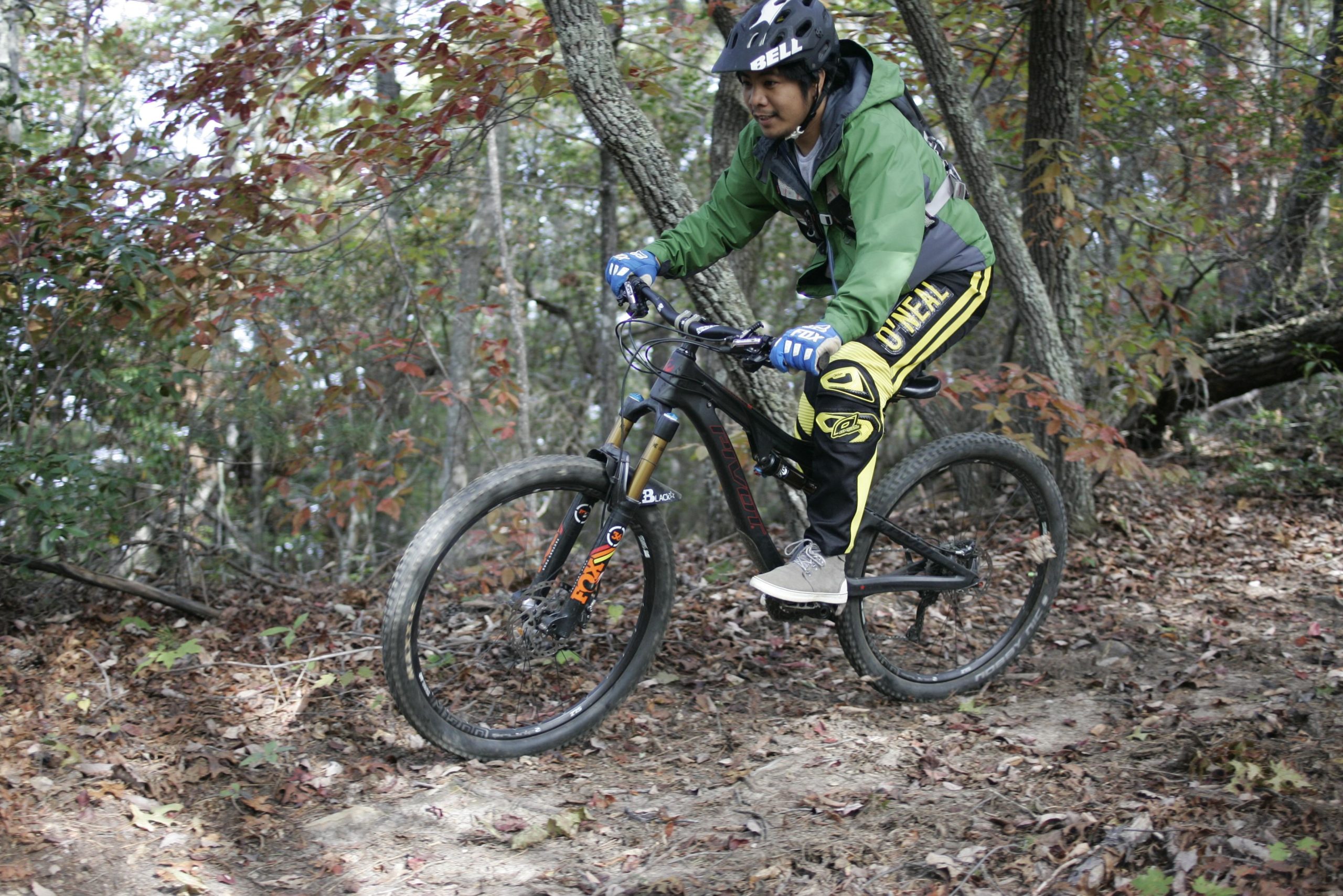 A person riding a mountain bike on a trail surrounded by trees with autumn foliage. The rider is wearing a helmet, gloves, and a green jacket, with yellow and black riding pants. The ground is covered with leaves, and the scene captures a dynamic moment of biking in nature. Sinkhole mountain bike trail.