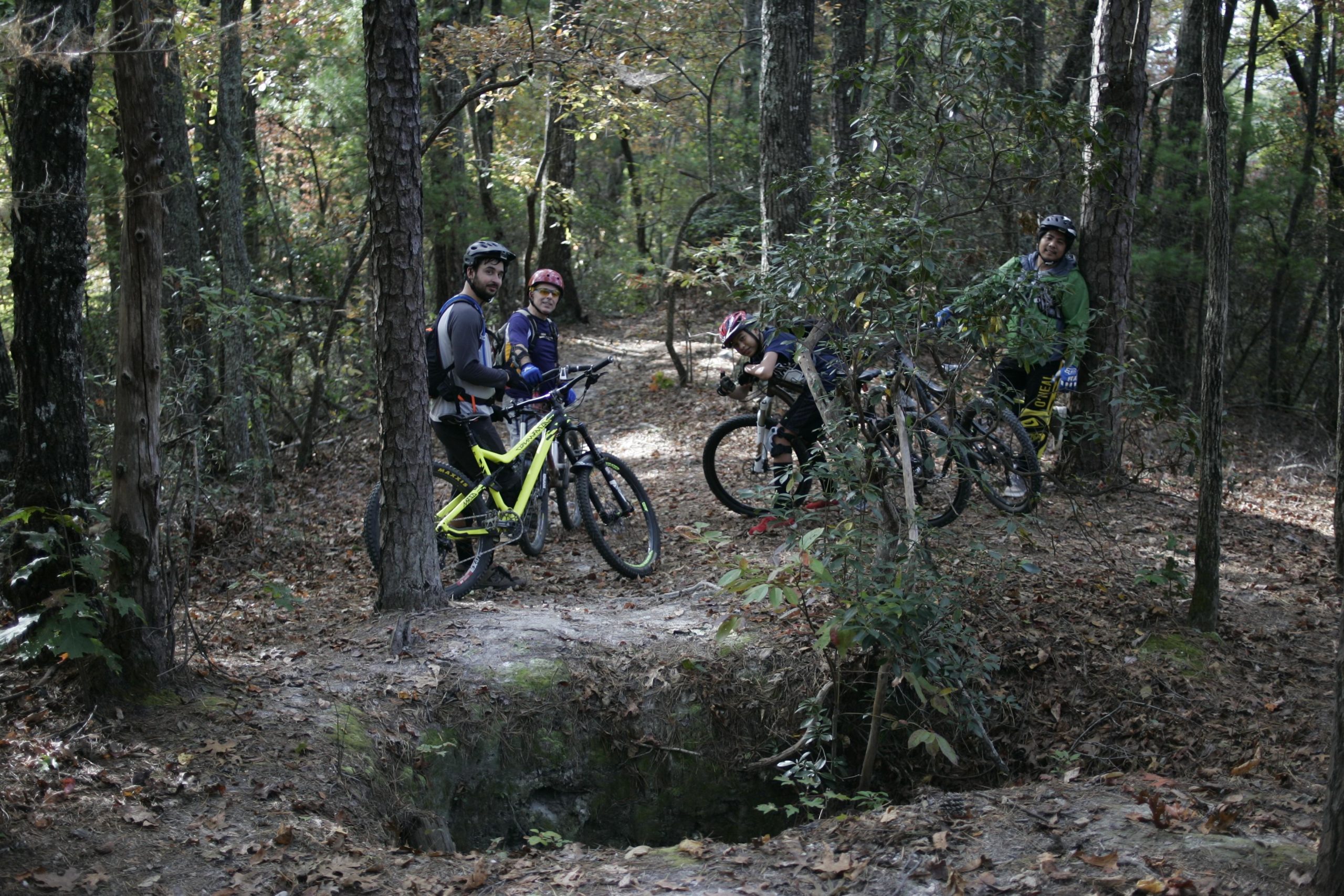 Four mountain bikers are gathered in a wooded area, surrounded by tall trees and fallen leaves. They are wearing helmets and cycling gear, with two bicycles leaning against trees. The scene captures a moment of camaraderie as they pause on their ride to chat and enjoy the natural surroundings. Sinkhole mountain bike trail.