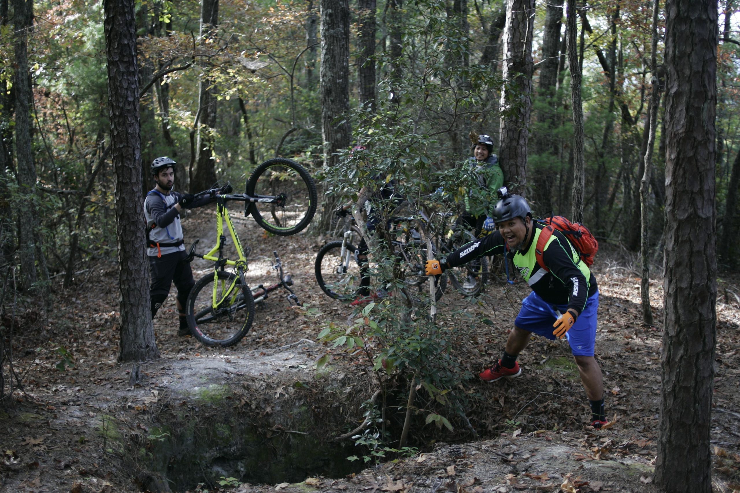 Four mountain bikers are captured in a wooded area, with one rider lifting their bike's front wheel off the ground, showcasing a trick. Another rider, in a bright green jersey, appears to be excitedly posing next to a small ravine. The scene is surrounded by trees and fallen leaves, indicating an autumn atmosphere. Sinkhole mountain bike trail.