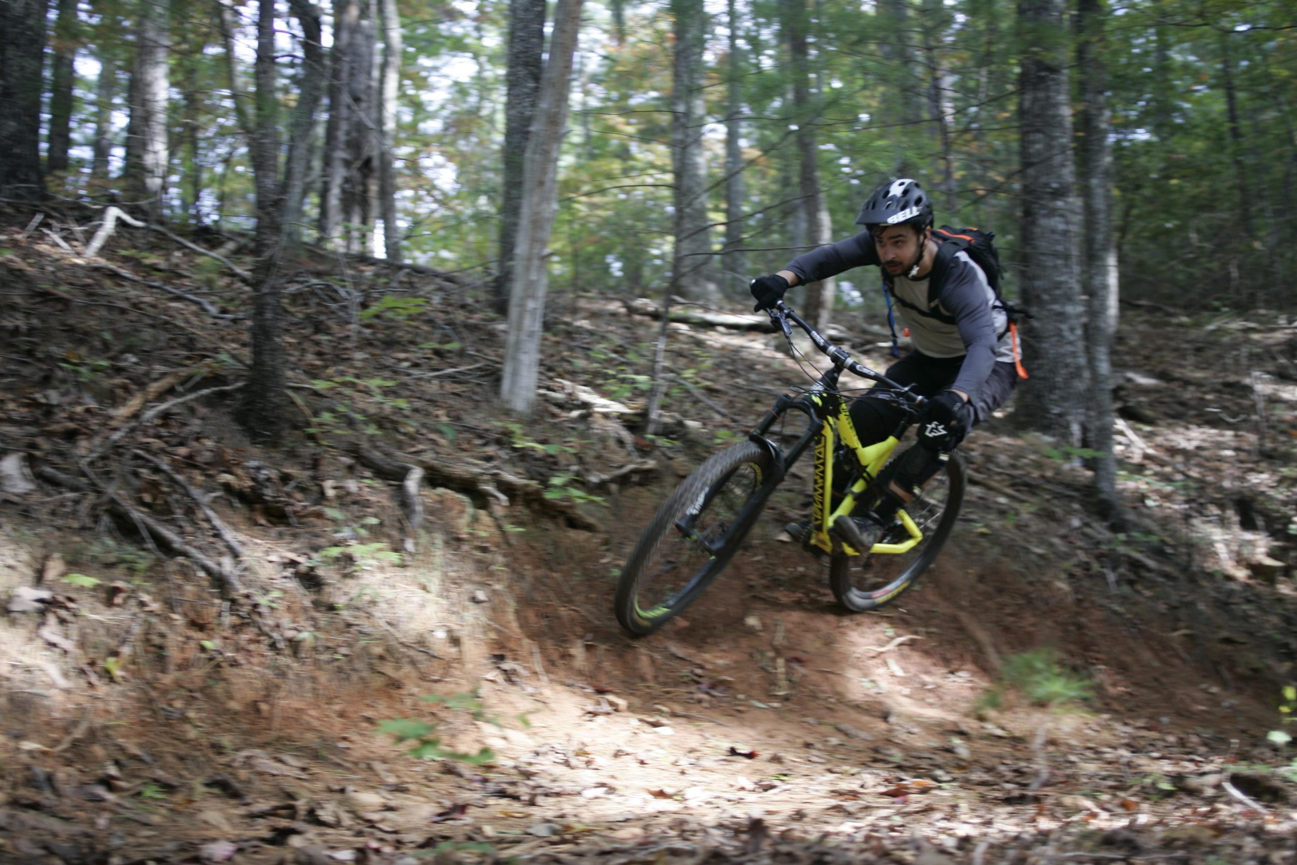 A mountain biker navigating a forest trail, leaning into a turn on a dirt path surrounded by trees and foliage. The cyclist wears a helmet and biking gear, showcasing an active outdoor lifestyle. Table Rock mountain bike trail.
