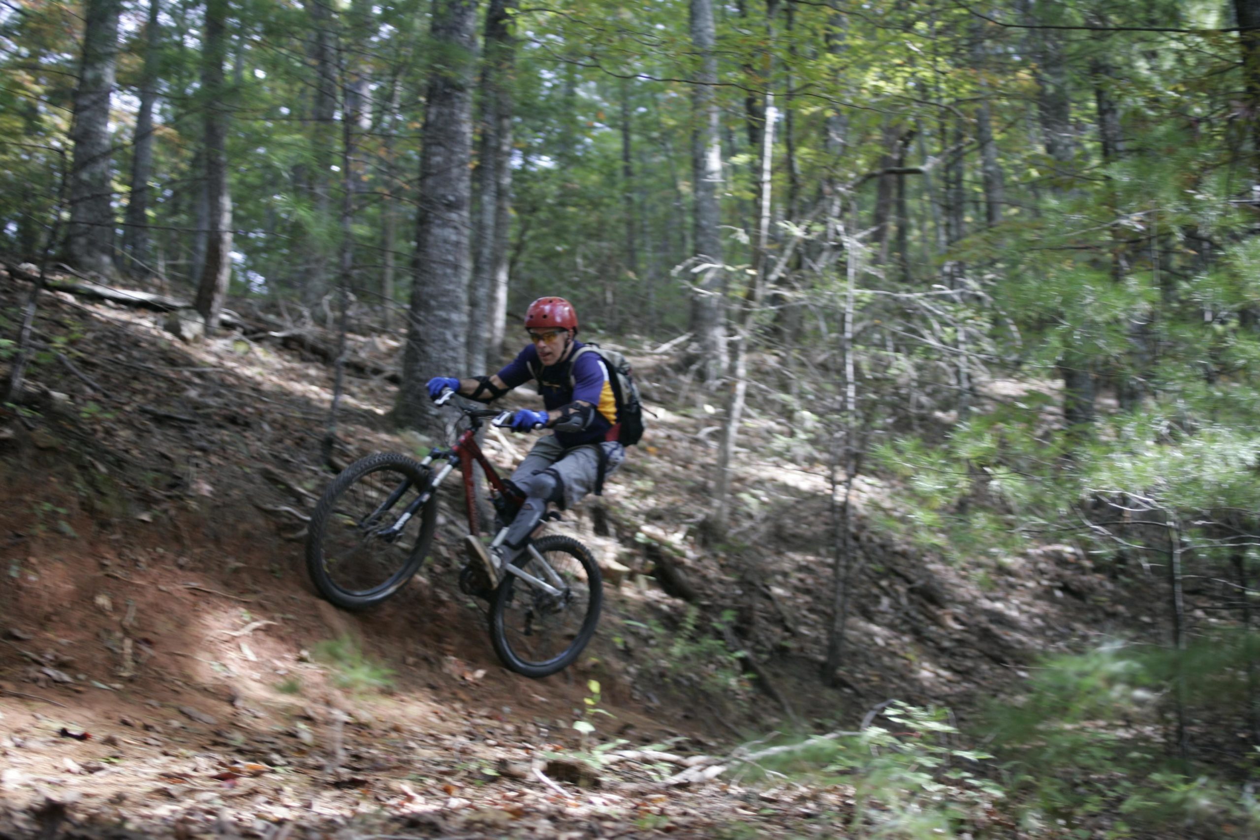 A mountain biker in a red helmet and blue gloves is jumping over a small ridge on a dirt trail surrounded by trees and foliage. The biker is wearing a backpack and gray pants, showcasing an action-packed moment in a forested area. Table Rock mountain bike trail.
