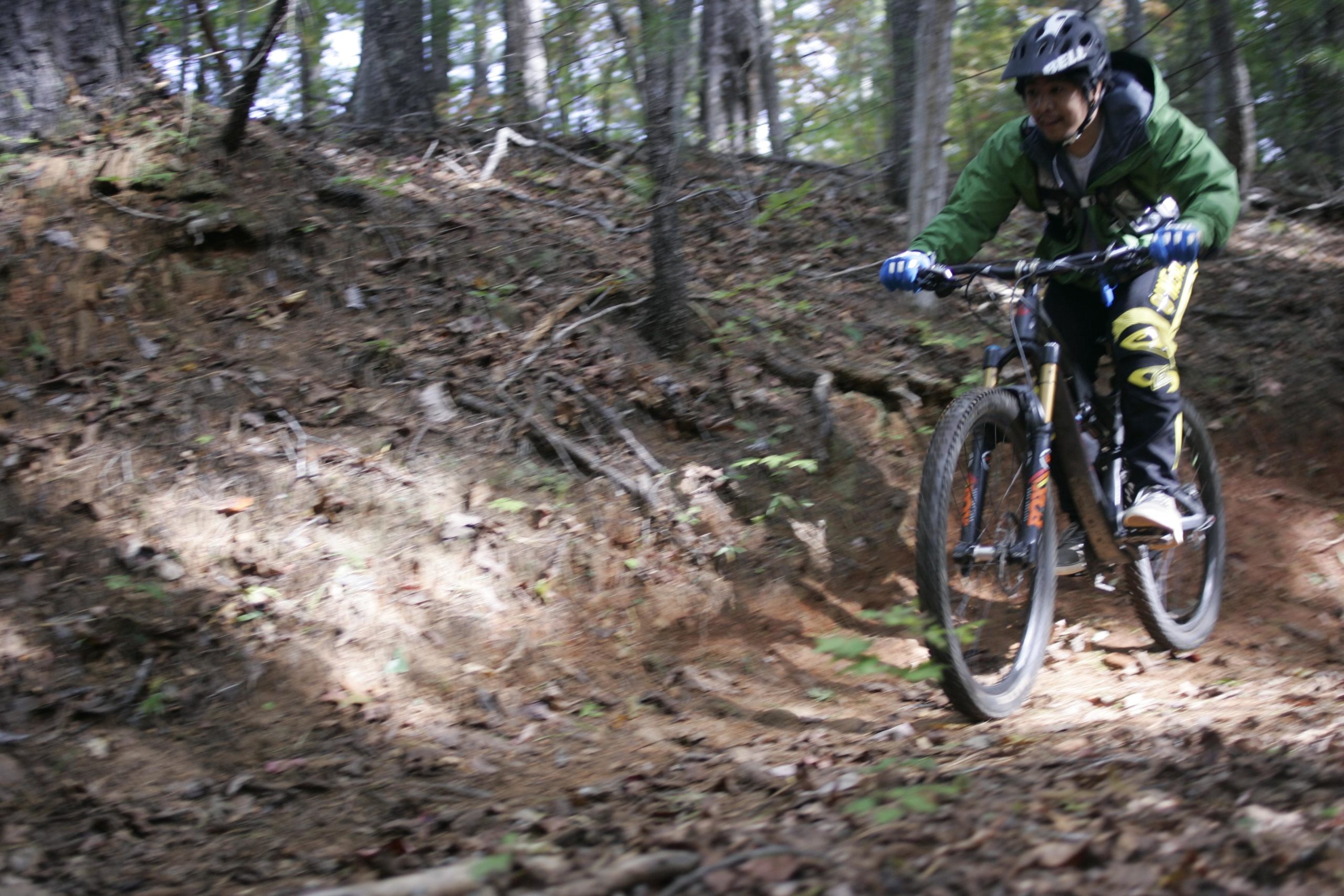 A mountain biker in a green jacket and helmet navigates a dirt trail surrounded by trees, with fallen leaves and roots visible on the ground. The rider is actively leaning forward on the bike, showcasing dynamic movement through the forested terrain. Table Rock mountain bike trail.