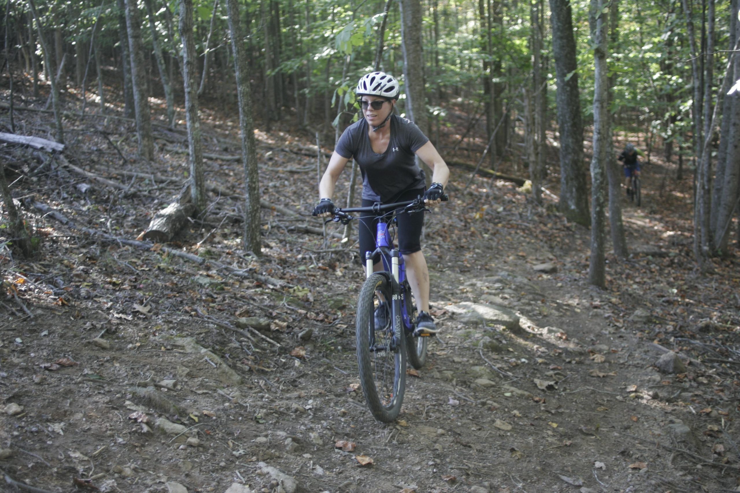 A woman riding a mountain bike on a rocky trail in a wooded area, wearing a helmet and sunglasses. Trees and fallen leaves surround the path, with a second biker visible in the background. Mountain Laurel Trails mountain bike trail.
