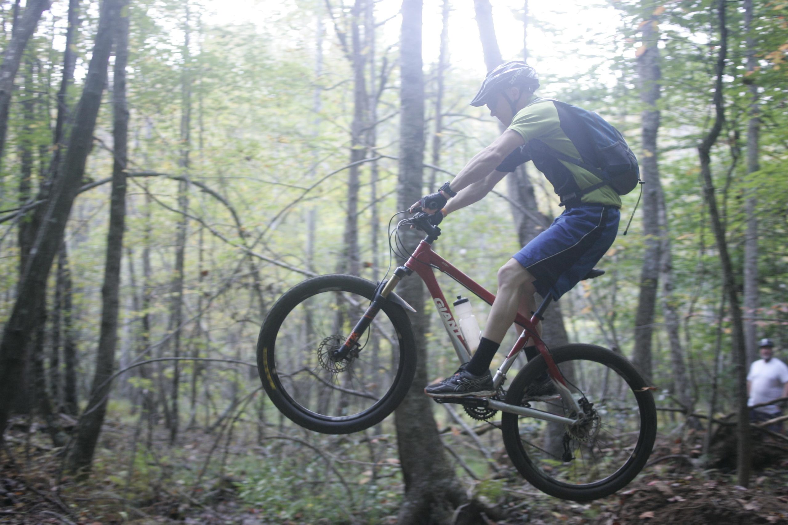 A mountain biker in a green and black jersey and blue shorts is captured mid-air as he jumps over a trail in a wooded area. He rides a red mountain bike and wears a helmet, with trees and a blurred figure in the background. The scene is set in a lush forest with dappled light filtering through the leaves. Mountain Laurel Trails mountain bike trail.