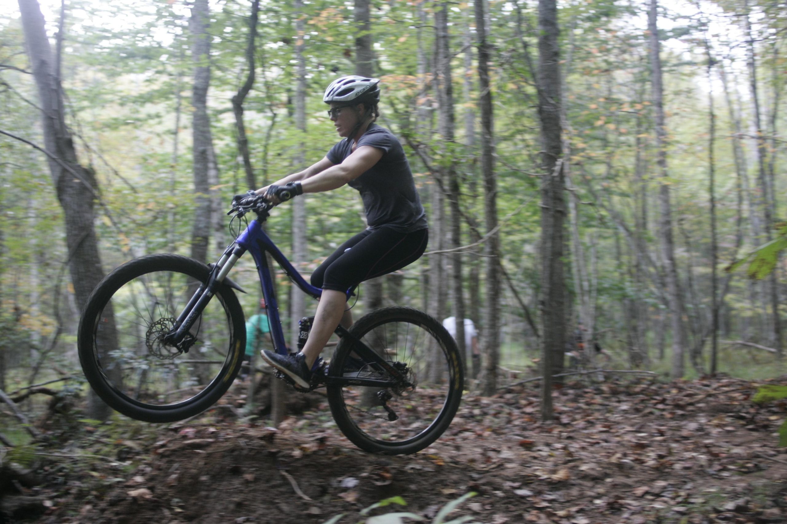 A mountain biker performing a wheelie on a dirt trail in a forested area, surrounded by trees and fallen leaves. The biker is wearing a helmet and athletic gear, demonstrating skill and balance while navigating the trail. Mountain Laurel Trails mountain bike trail.
