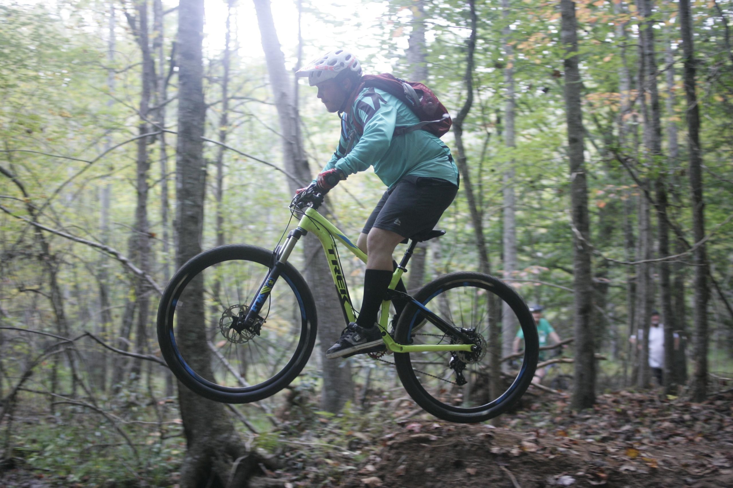 A mountain biker in a teal jersey and helmet performs a jump on a trail, surrounded by trees and foliage. The bike's front wheel is raised off the ground, showcasing an adventurous moment in nature. Mountain Laurel Trails mountain bike trail.