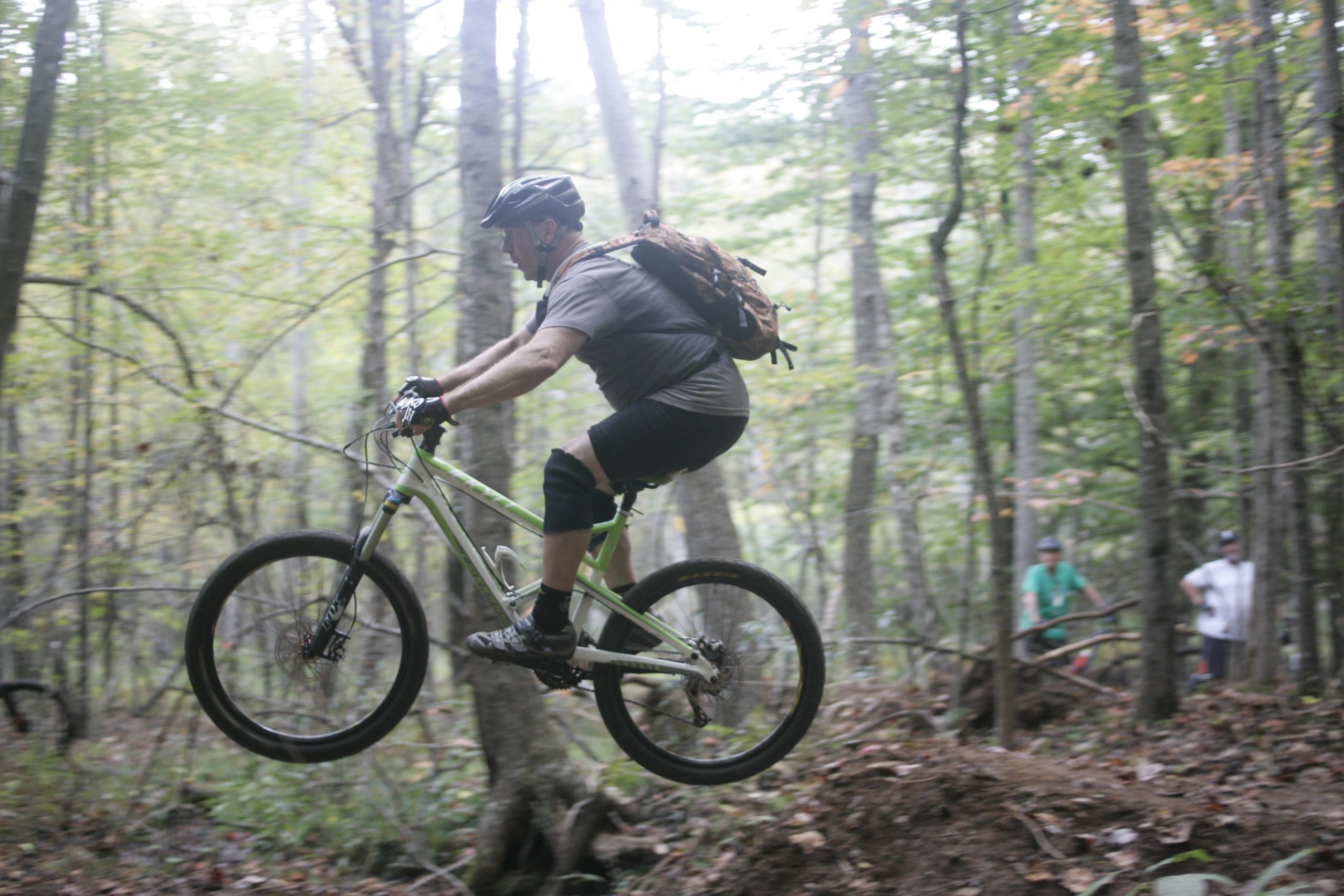 A mountain biker performing a jump on a trail in a wooded area, with trees and foliage surrounding the scene. The rider is wearing a helmet and protective gear, showcasing an action-packed moment in nature. In the background, a few spectators can be seen watching the jump. Mountain Laurel Trails mountain bike trail.