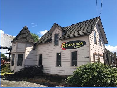 A pink house with a unique architectural design, featuring turrets and a sloped roof, displaying a sign that reads "Revolution Cycles." The building is surrounded by greenery and set against a clear blue sky. The street number "3506" is visible on the front of the house.