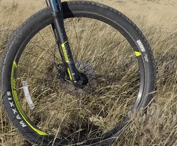 Maxxis Ikon: Close-up of a mountain bike front tire resting on tall grass, featuring a black wheel rim and a yellow-highlighted tire design.