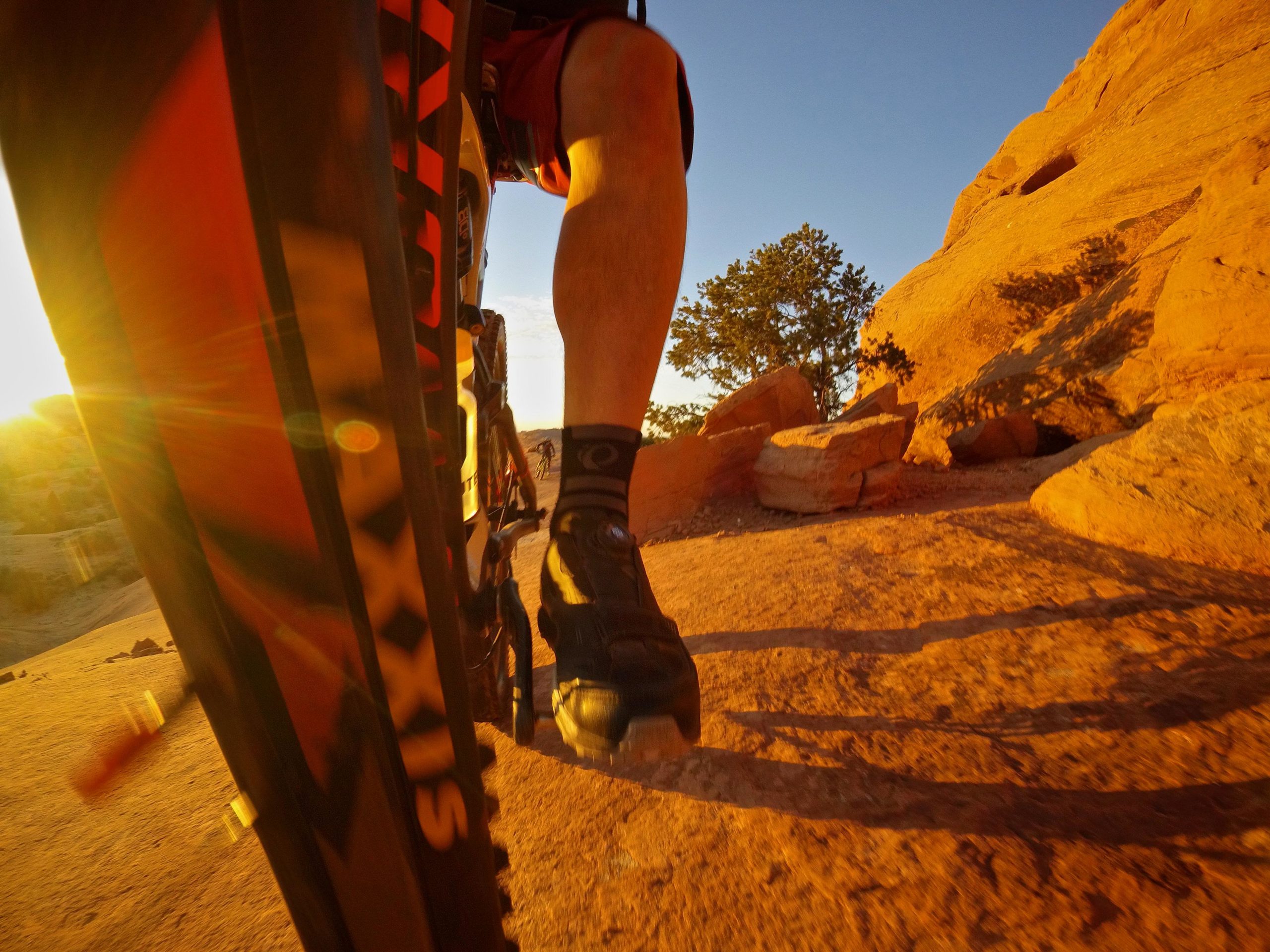 A close-up view of a mountain bike rider's leg and wheel on a rocky trail during sunset, with warm light illuminating the landscape and shadows cast on the ground. Slickrock mountain bike trail.